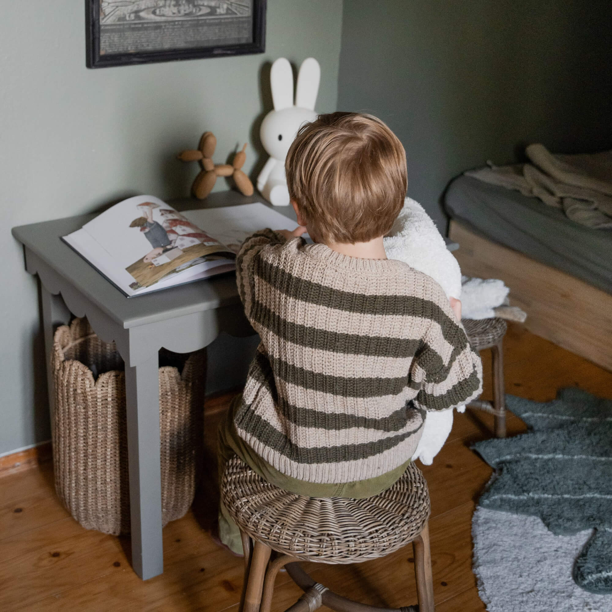 Kind im beige-grünen Pullover sitzt auf Rattanhocker an grauem Holztisch mit Buch und Hasenlampe