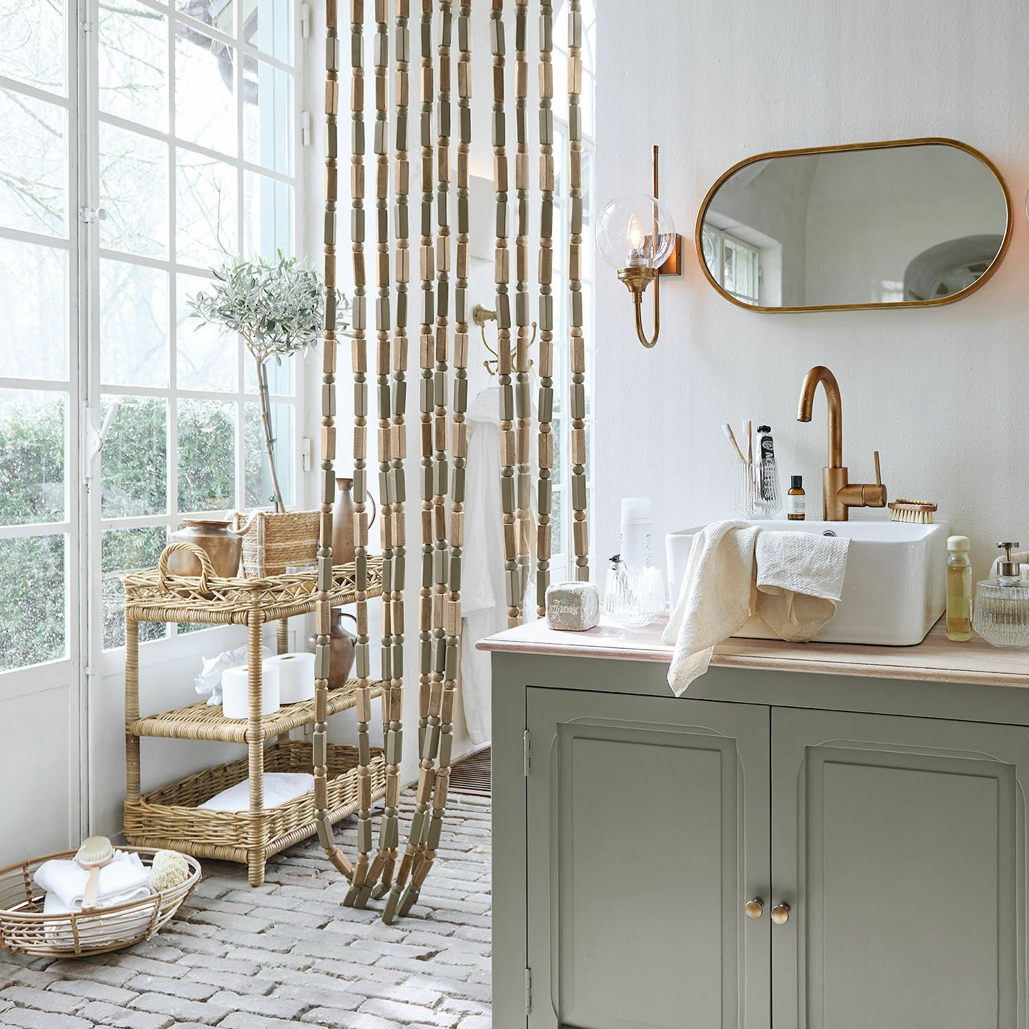 Badezimmer mit grünem Schrank, weißem Waschbecken, goldfarbenem Wasserhahn, natürlichem Holzparavent und Rattanständern