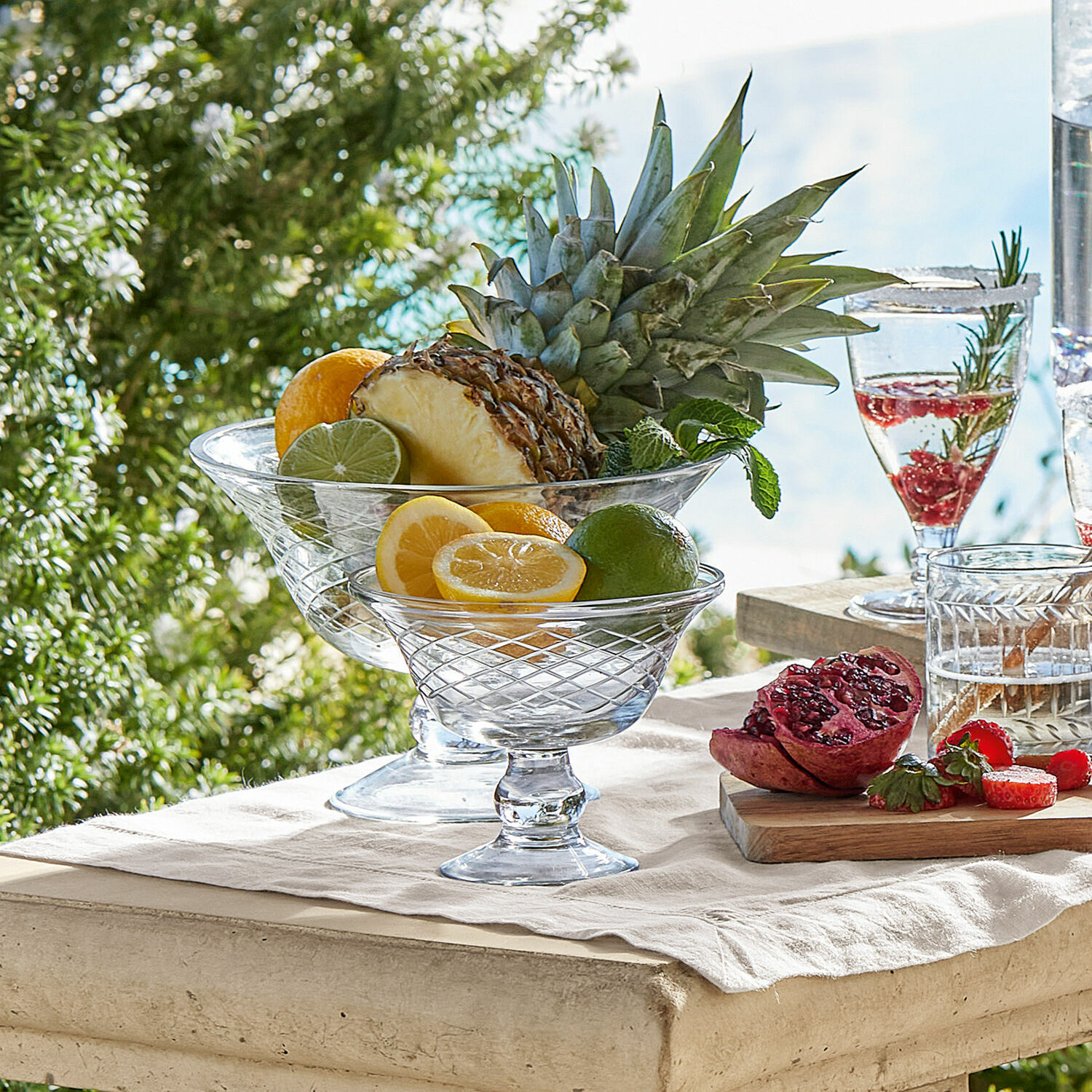 Glas-Schalen auf Fuß mit Zitrusfrüchten und Ananas auf hellem Tisch im Freien