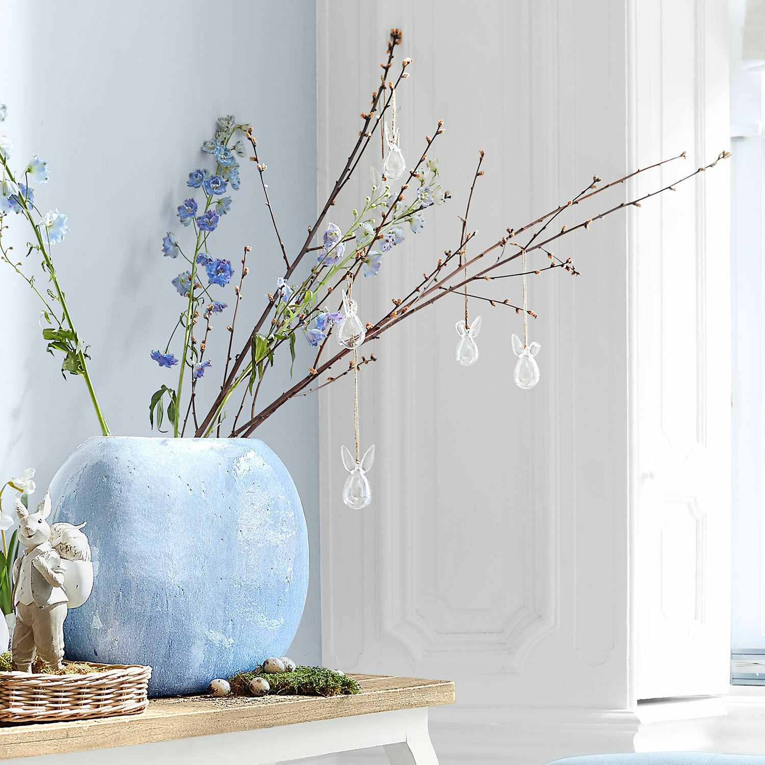 Blaue Keramikvase mit Zweigen und gläsernen Hasenanhängern auf Holztisch dekoriert