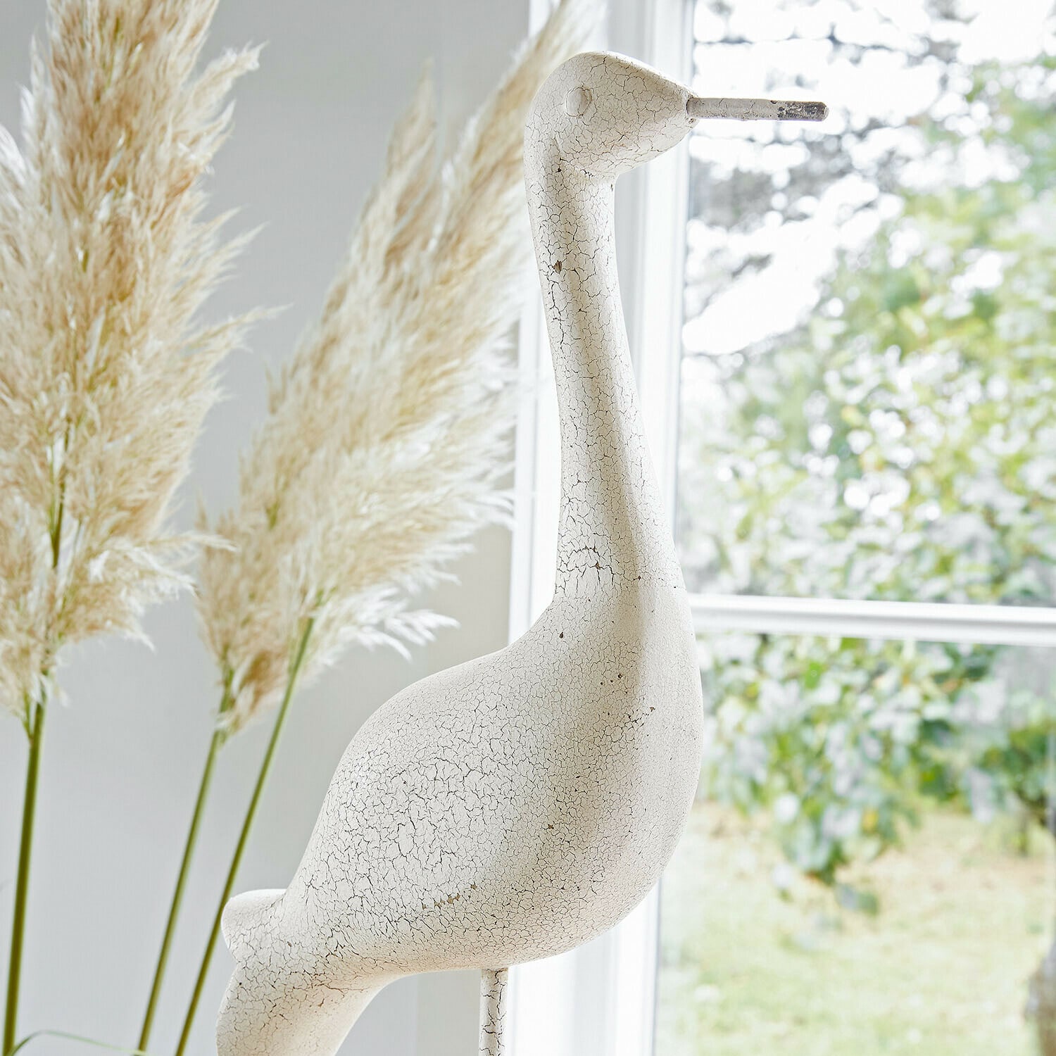 Weiße, rissige Vogel-Dekoration aus Keramik vor Fenster mit Pampasgras im Hintergrund