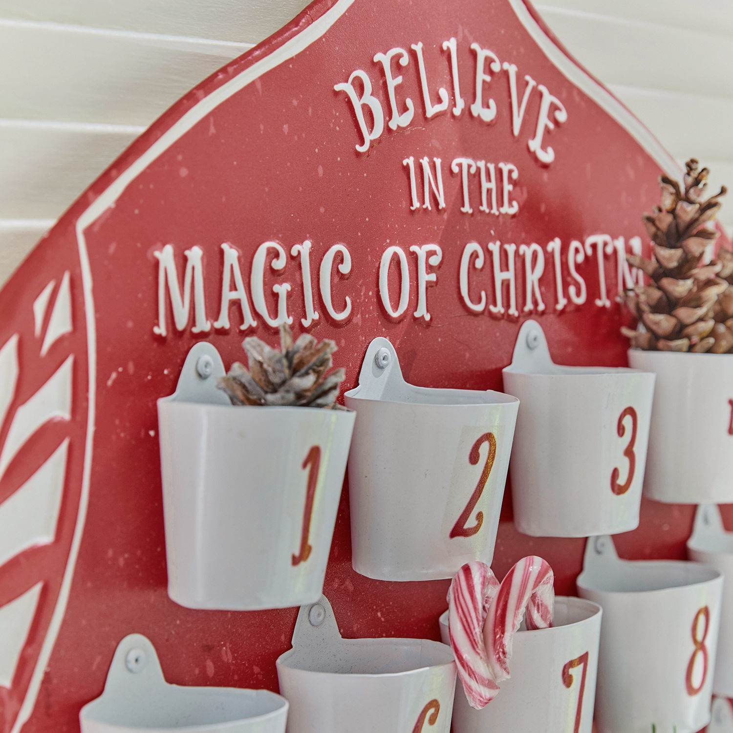 Roter Adventskalender mit weißen Metallbechern und Weihnachtsdekorationen aus Zapfen und Zuckerstangen