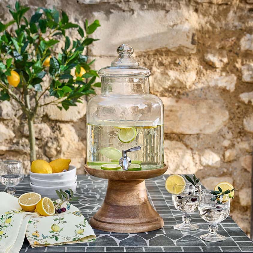 Glasgetränkespender mit Deckel auf Holzsockel, Limetten- und Zitronenscheiben auf dem Tisch
