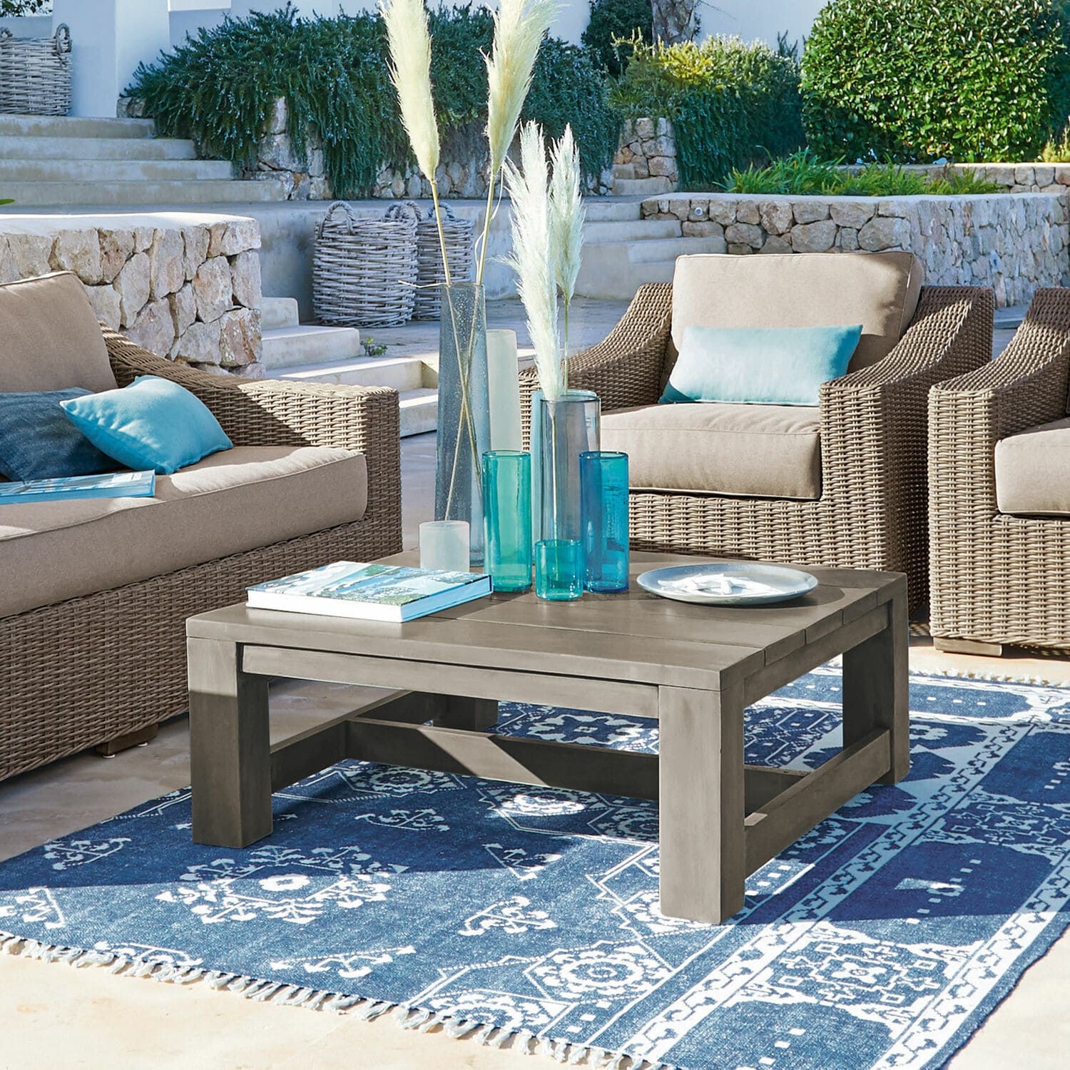 Grauer Holzcouchtisch auf blauem gemustertem Teppich, umgeben von Korbsesseln mit blauen Kissen.