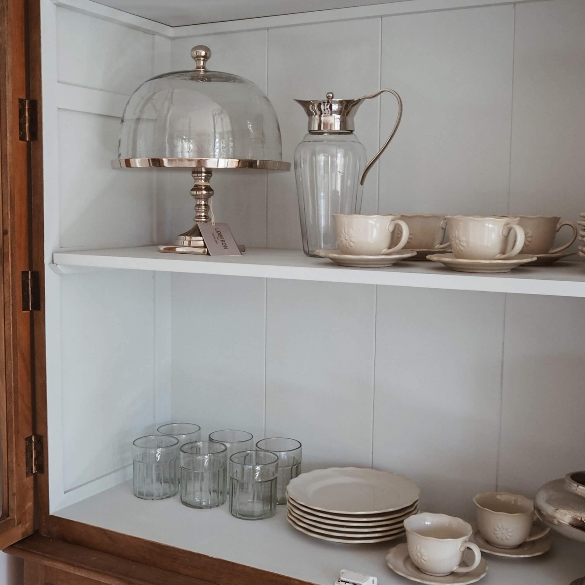 Set aus cremefarbenen Keramiktassen und Tellern mit Glas-Krug und Kuchenhaube im weißen Schrank