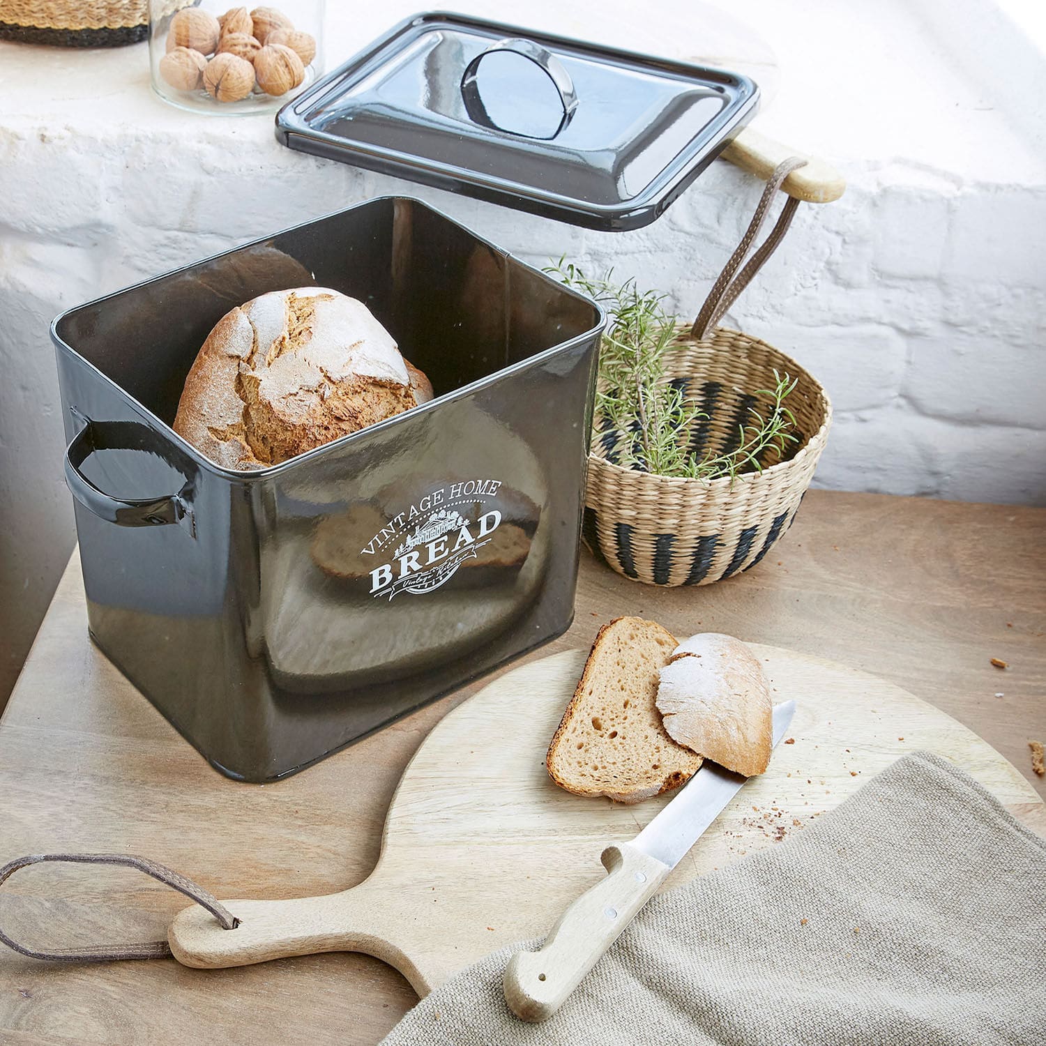 Schwarzer Metall-Brotkasten mit Deckel und frischem Brot auf Holzschneidebrett
