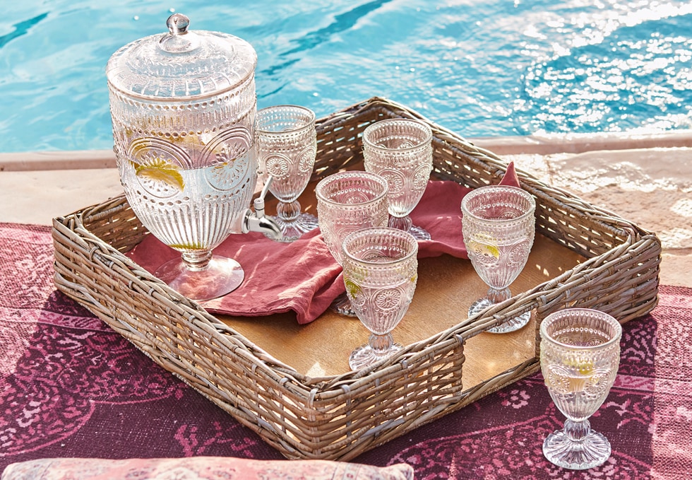 Glas-Krug und sechs verzierte Gläser auf einem Weidenkorb-Tablett am Pool, sommerliches Getränkeset