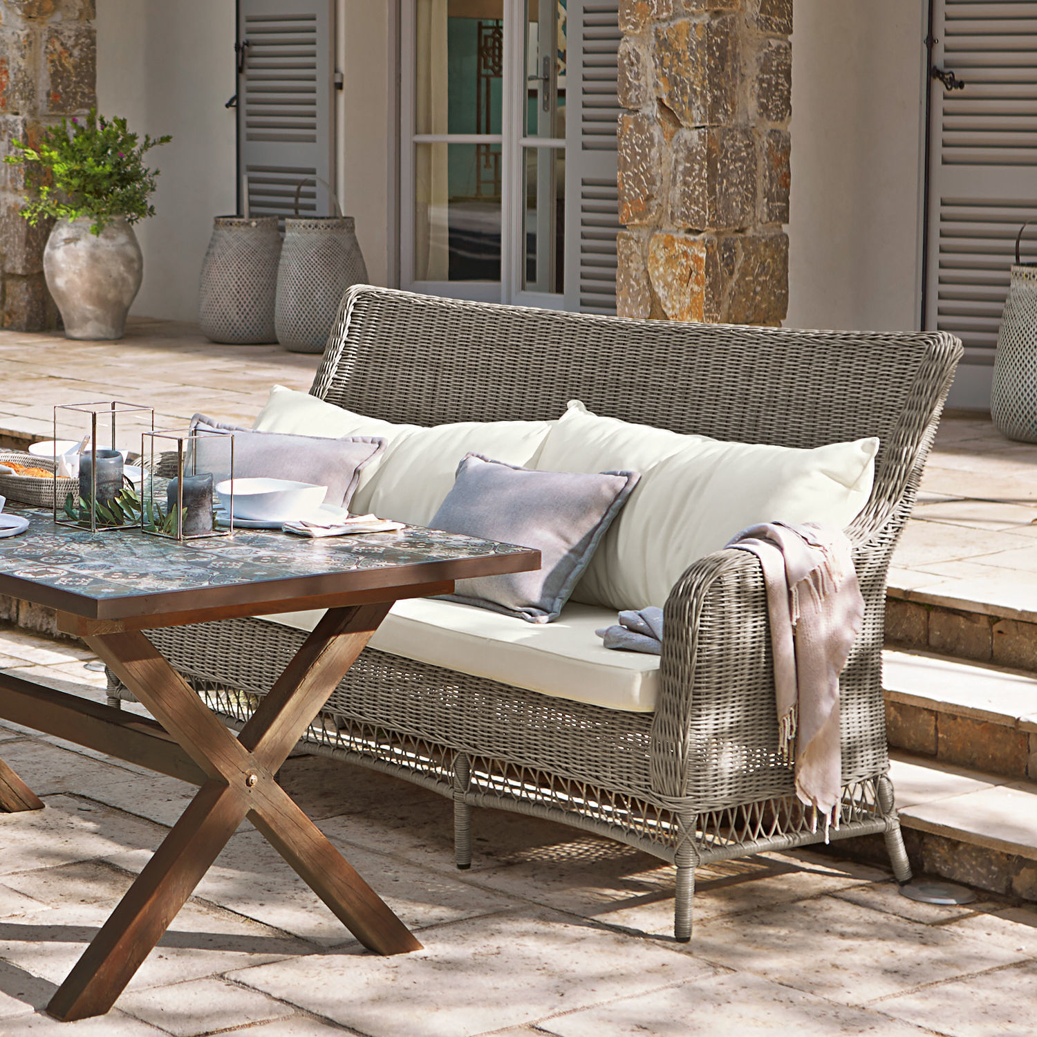 Graues Rattan-Sofa mit cremefarbenen Kissen auf Terrasse mit Holztisch und Dekorationen