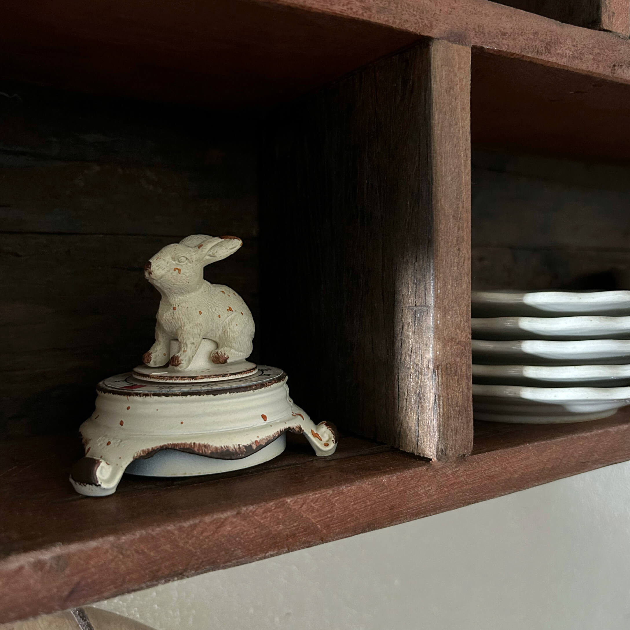 Weißer Keramikbehälter mit Hasenfigur als Deckel auf Holzregal neben Tellern