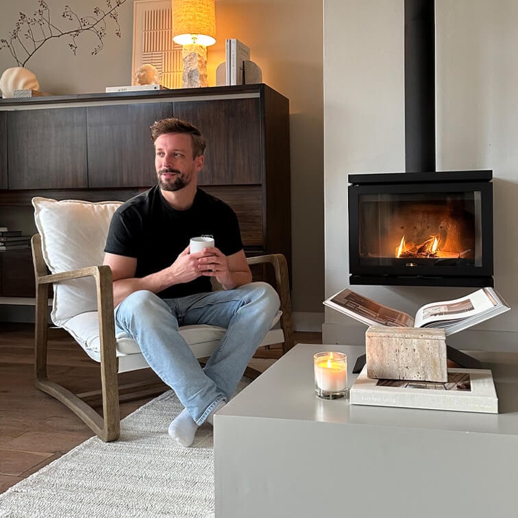 Mann sitzt auf Holzstuhl mit weißem Kissen, hält Tasse, Kamin und Kerze auf dem Tisch im Wohnzimmer