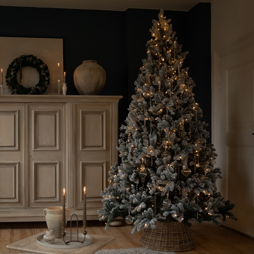 Künstlicher, schneebedeckter Weihnachtsbaum mit warmen Lichtern und Dekoration in gemütlichem Raum