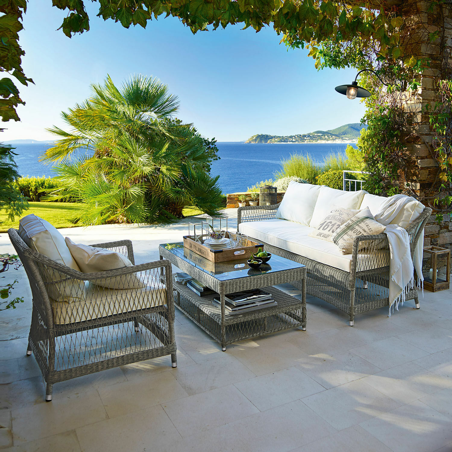 Gartenmöbel-Set aus grauem Rattan mit weißen Kissen auf Terrasse mit Meerblick