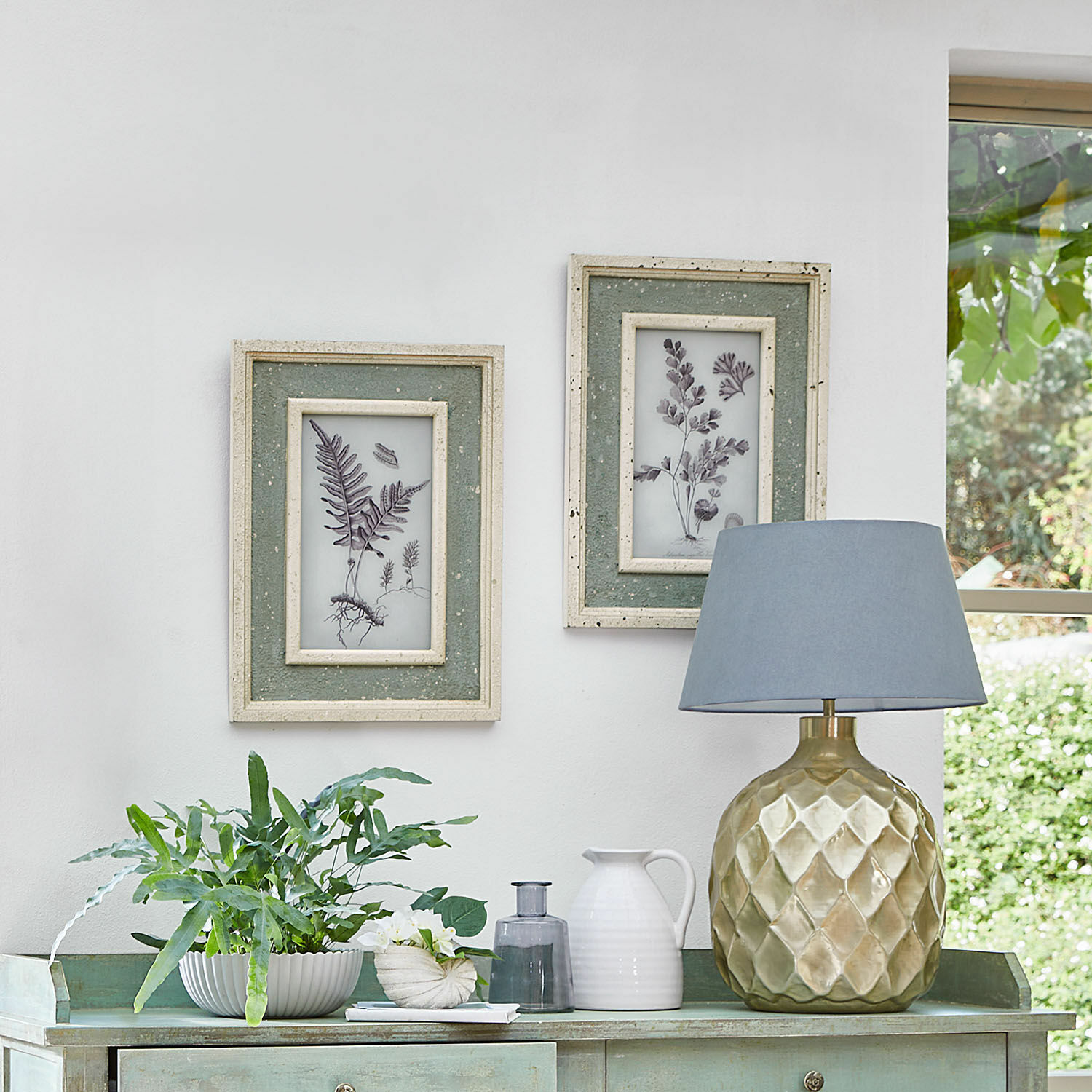 Goldene Keramiklampe mit blauem Lampenschirm auf grünem Sideboard mit Pflanzen und botanischen Bildern.