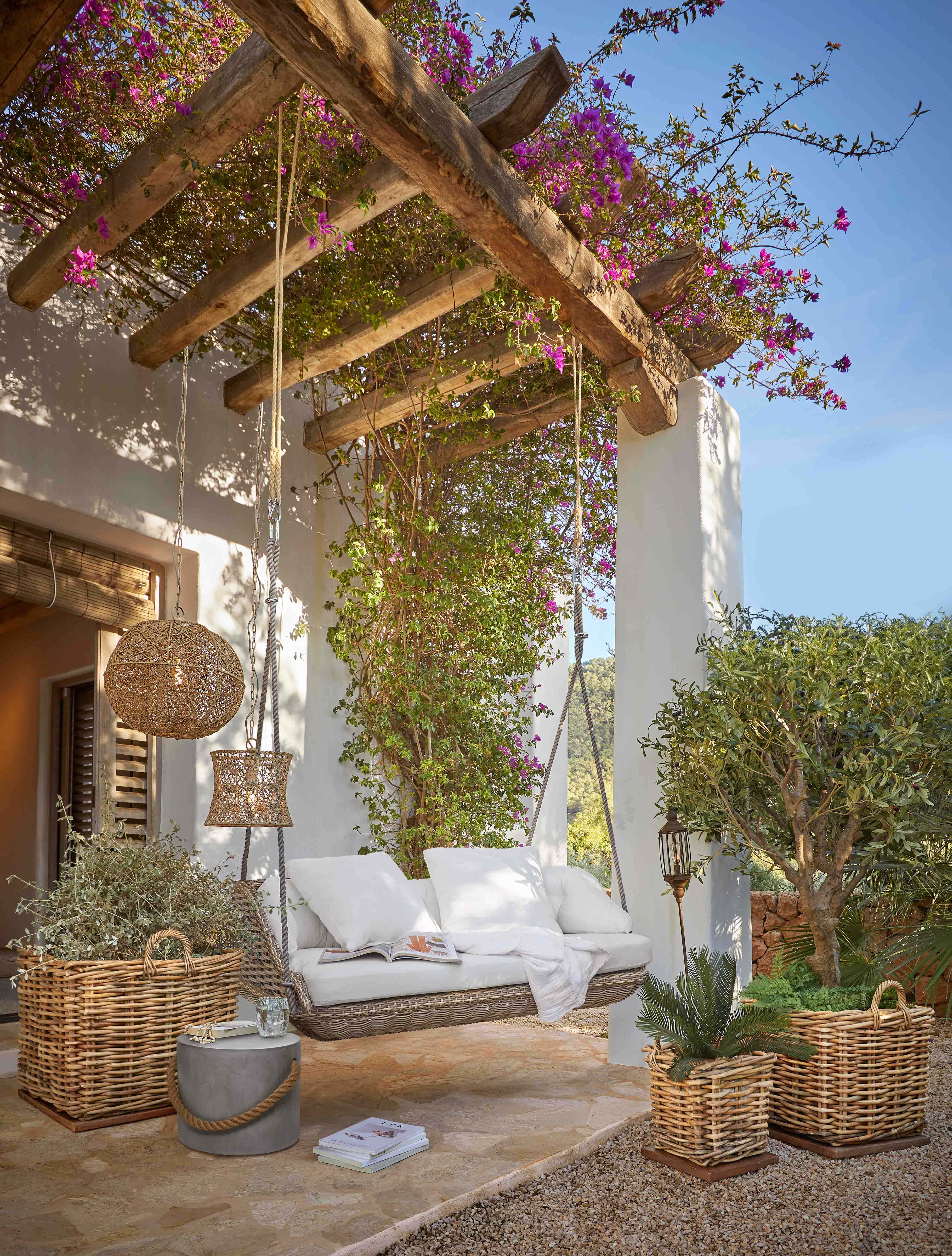 Wiklingschaukel mit weißen Kissen unter einer Holzpergola mit blühenden pinken Blumen