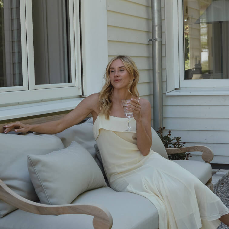 Frau im cremefarbenen Kleid sitzt entspannt auf hellgrauem Sofa draußen und hält ein Weinglas