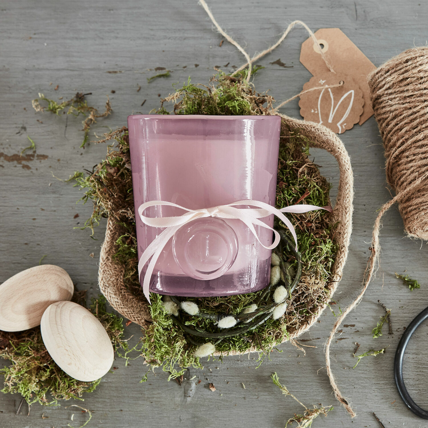 Violetter Glas-Teelichthalter mit weißer Schleife auf natürlichem Moos in Jutekorb, Dekoration