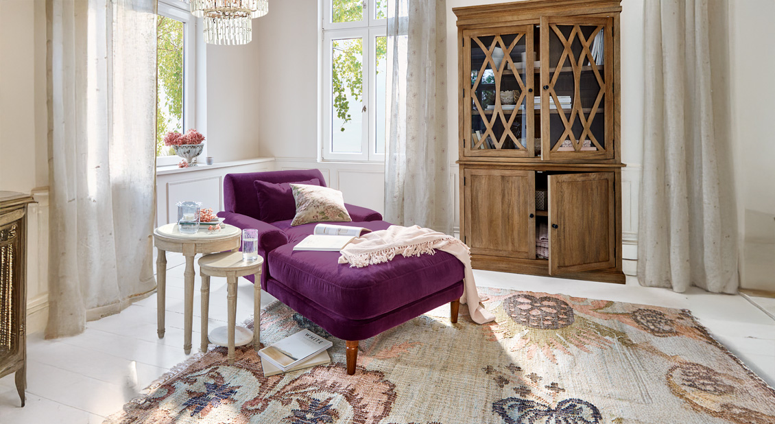 Violette Samt-Chaiselongue mit Kissen und Decke in hellem Wohnzimmer mit Holzschrank und Teppich