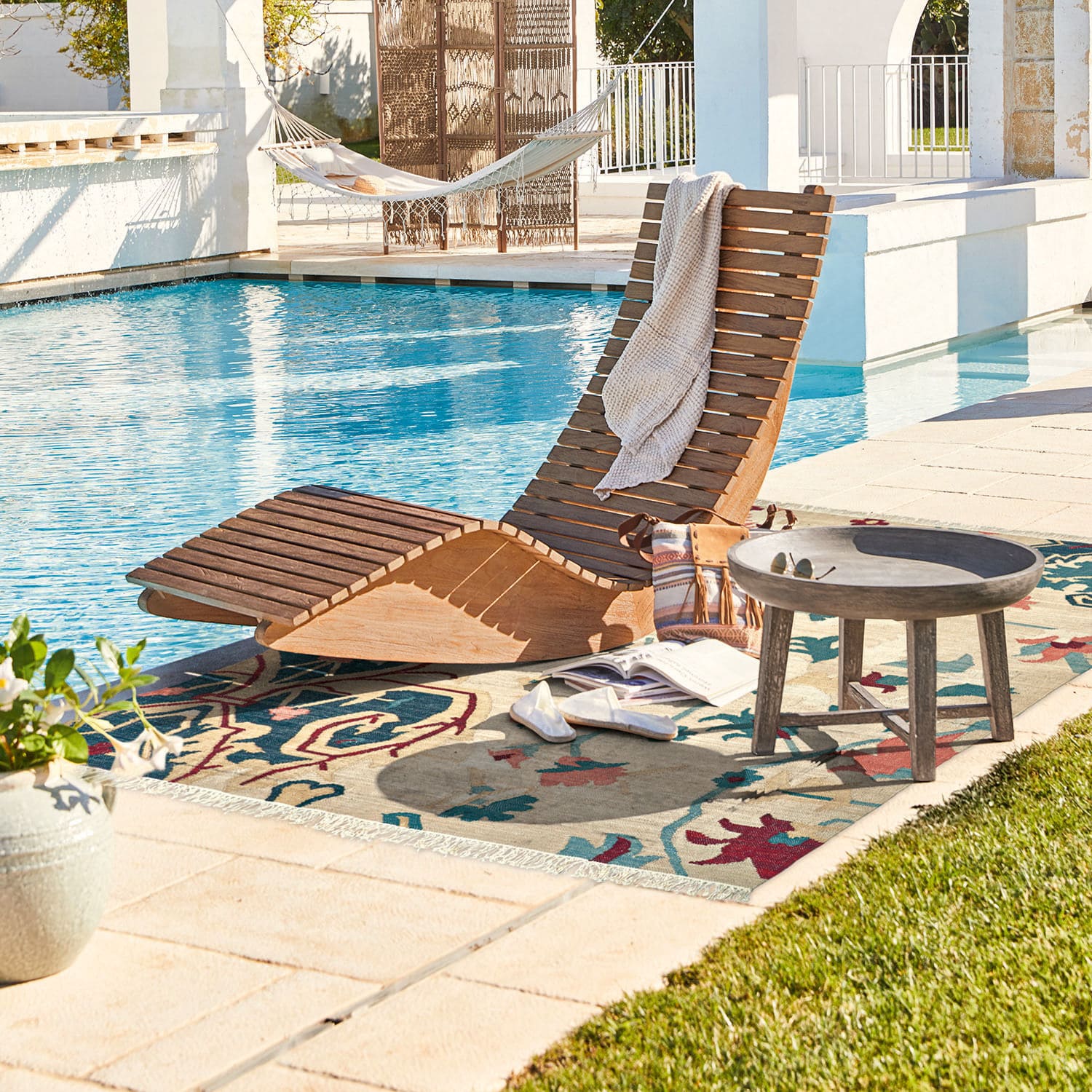 Holzliege in Naturfarbe am Pool mit buntem Teppich und rundem Beistelltisch aus Holz
