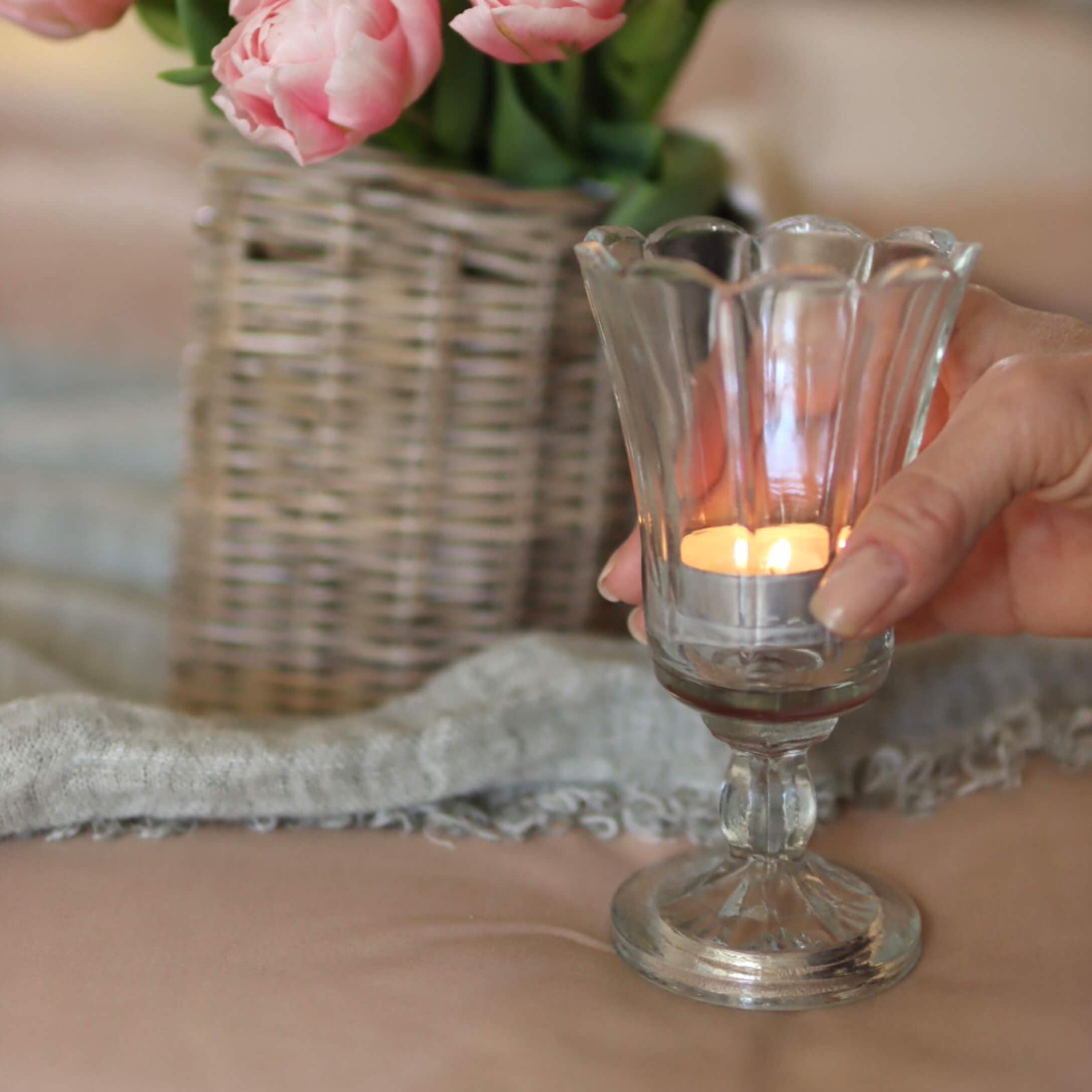 Glas-Teelichthalter auf Fuß mit brennender Kerze, rosa Blumen in Korbvase im Hintergrund