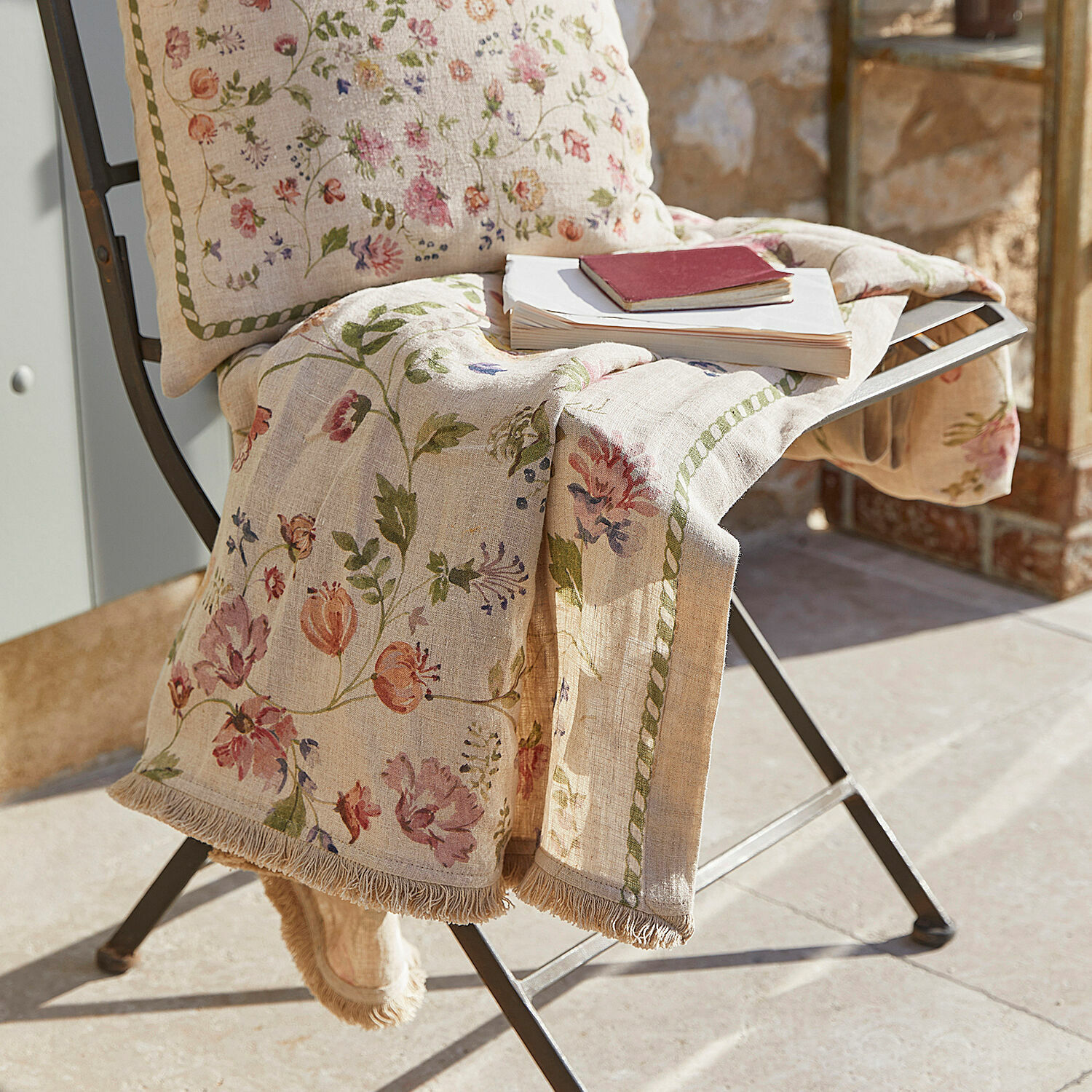 Beige Leinen-Decke mit floralem Muster und Fransen auf Metallstuhl im Sonnenlicht