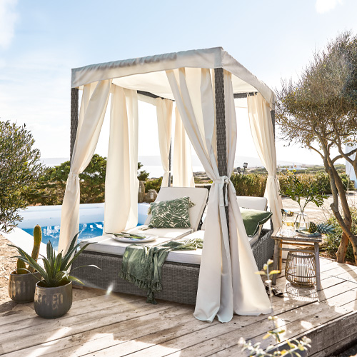 Graues Rattan-Liegebett mit cremefarbenen Vorhängen und grünen Kissen am Pool auf Holzterrasse