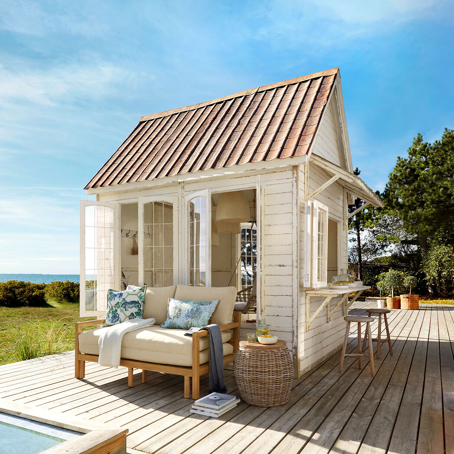Gartenhaus Stormhavn Weißes Holzhaus mit rotem Dach und beigem Sofa auf Holzterrasse mit Meerblick