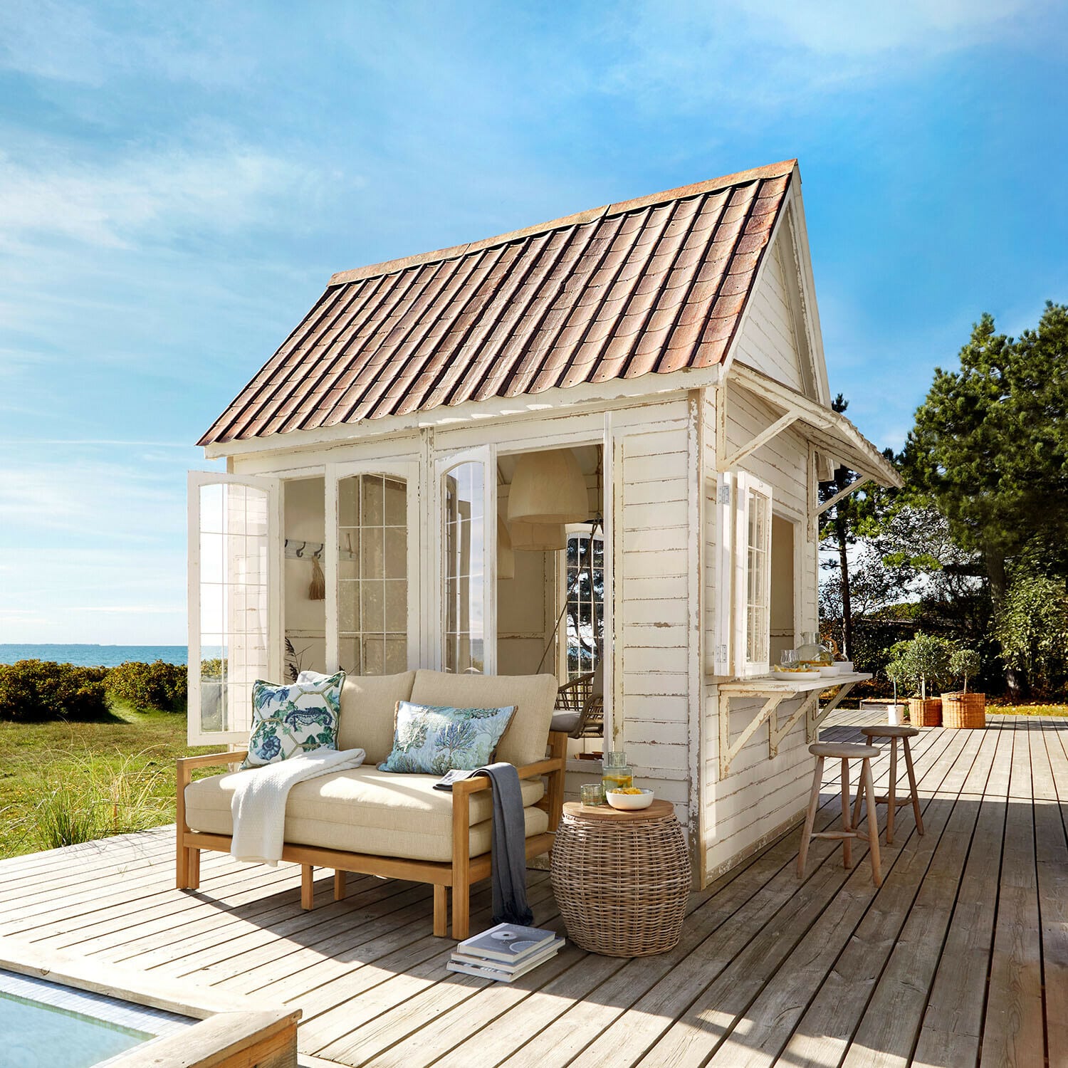 Gartenhaus Stormhavn Weißes Holzhaus mit rotem Dach und beigem Sofa auf Holzterrasse mit Meerblick