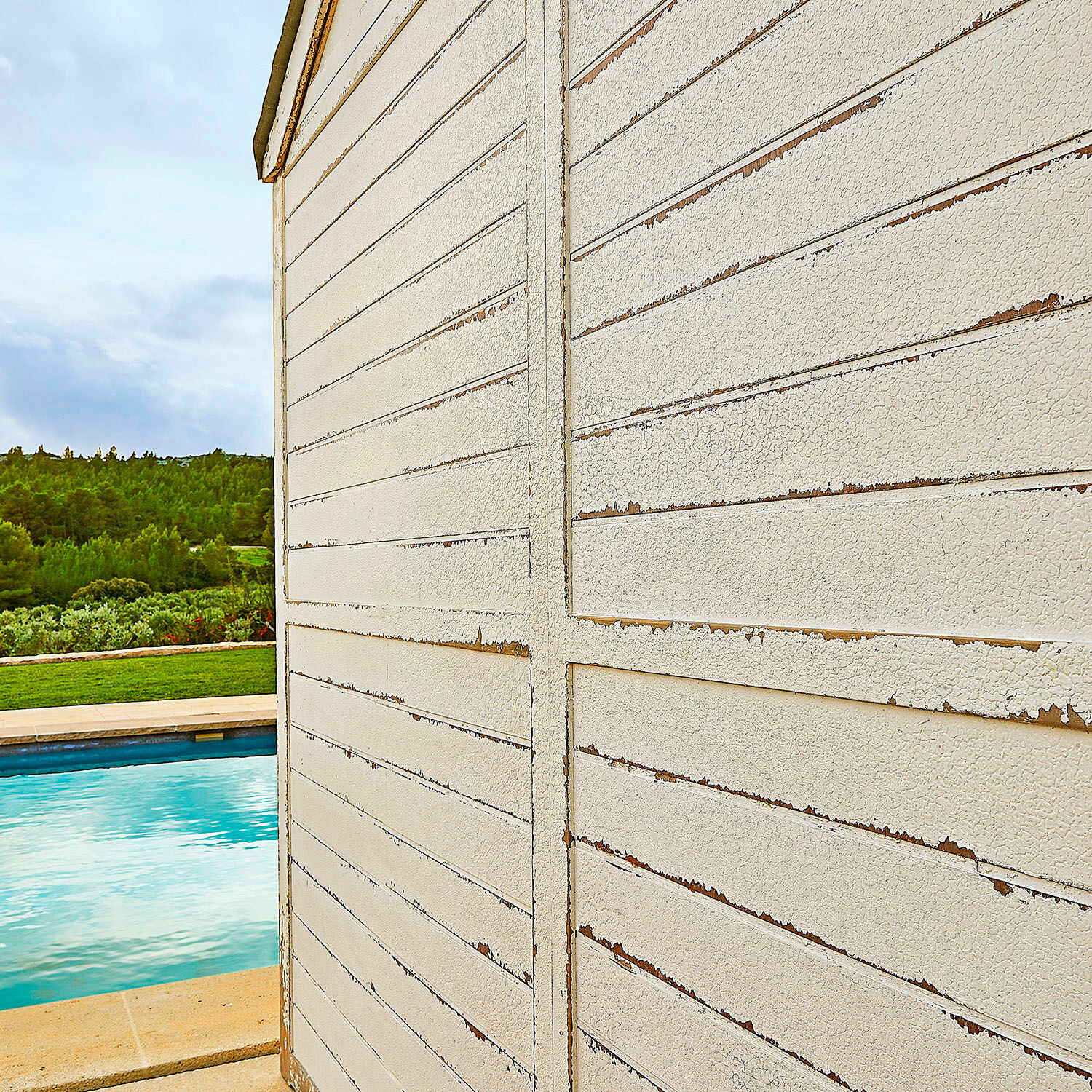 Weiße Holzfassade mit abblätternder Farbe außen neben Pool und grüner Landschaft