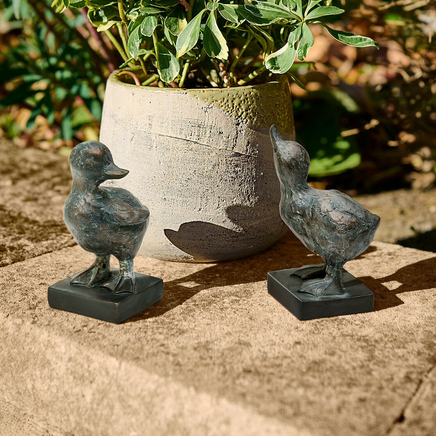 Zwei dunkle Metall-Entenfiguren auf Steinplatte neben einer Pflanzvase im Garten