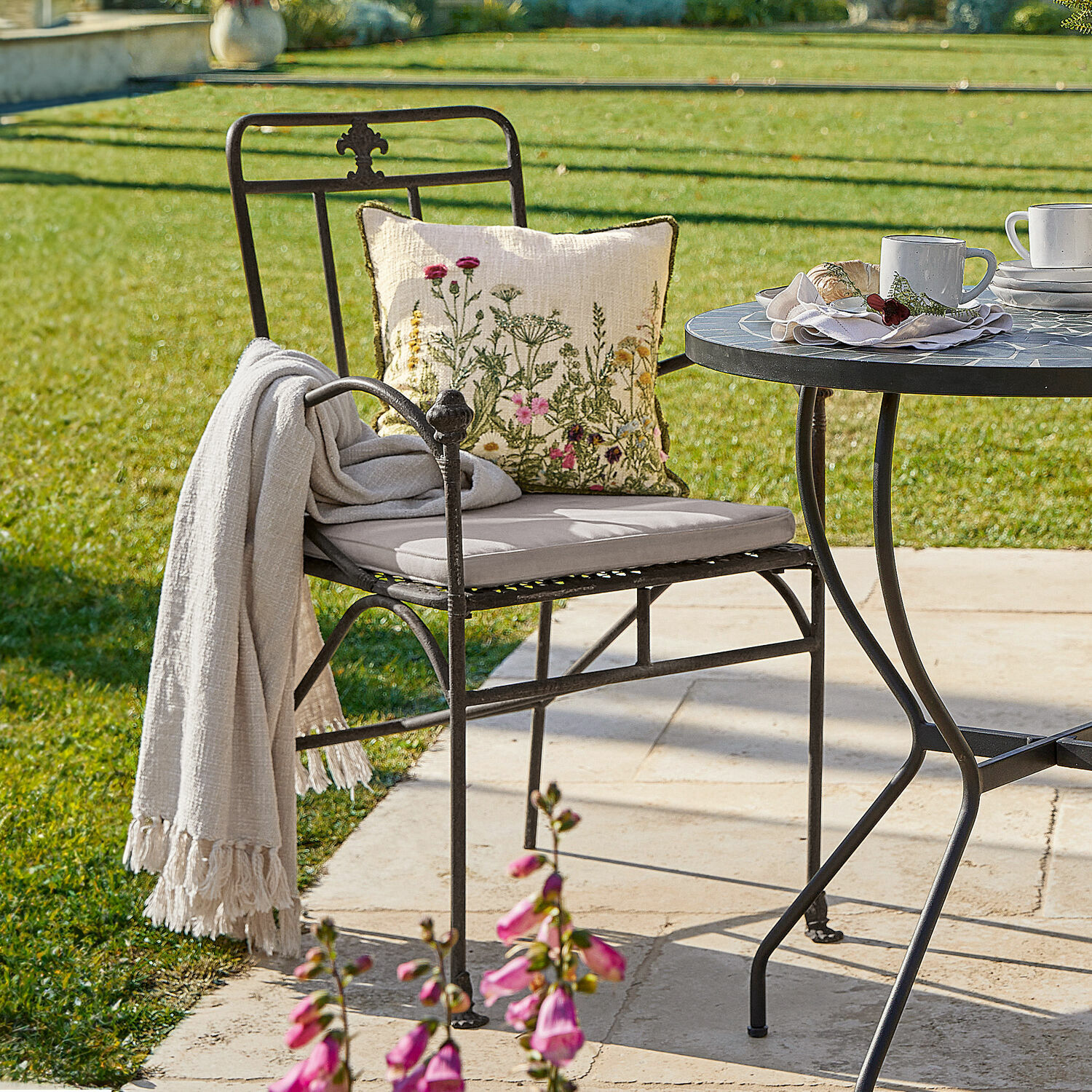 Stuhl Maidis Schwarzer Metall-Gartenstuhl mit hellem Sitzkissen und floralem Dekokissen auf sonniger Terrasse