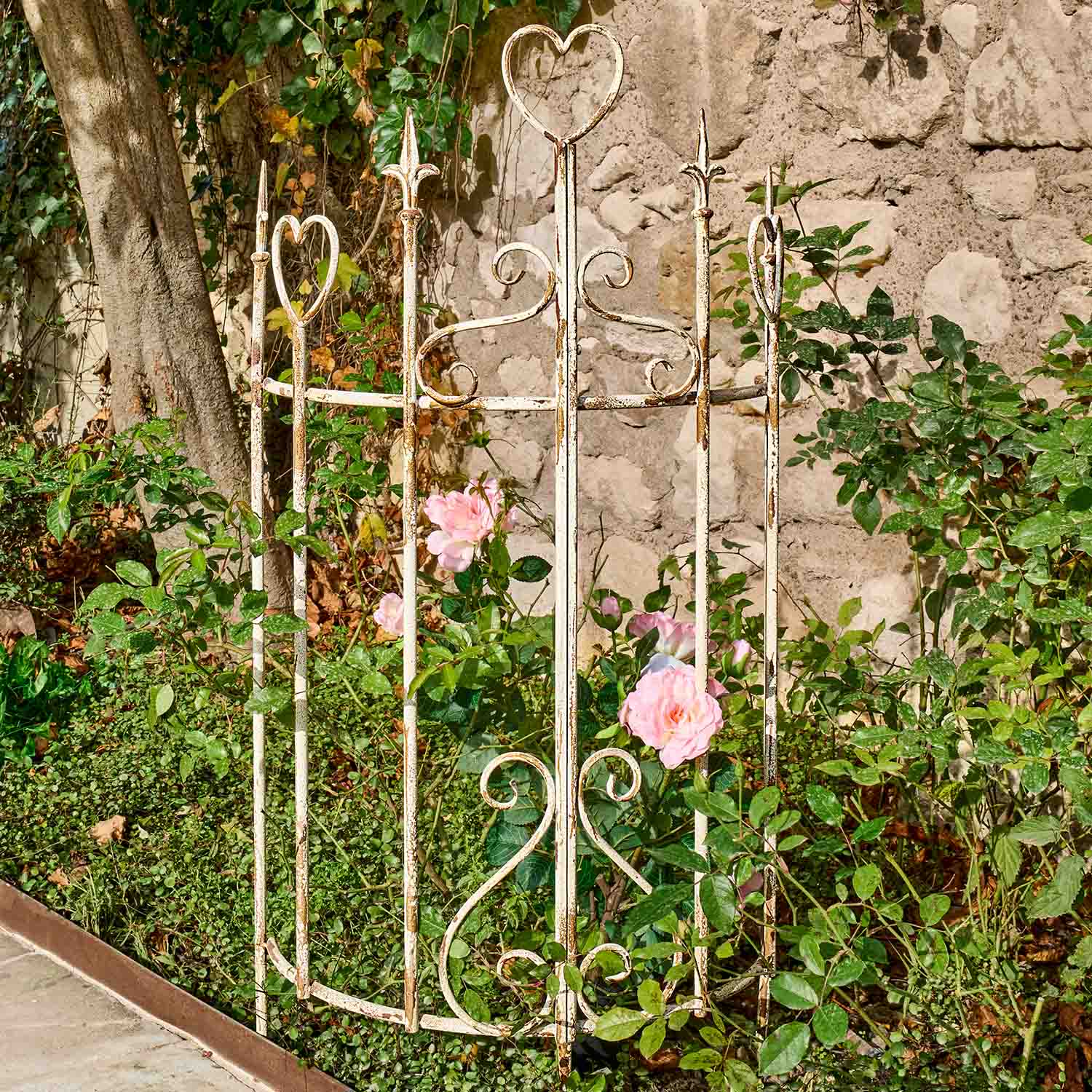 Weißes Metall-Gartengitter mit Herzmotiven und rosa Rosen im Garten vor Steinmauer