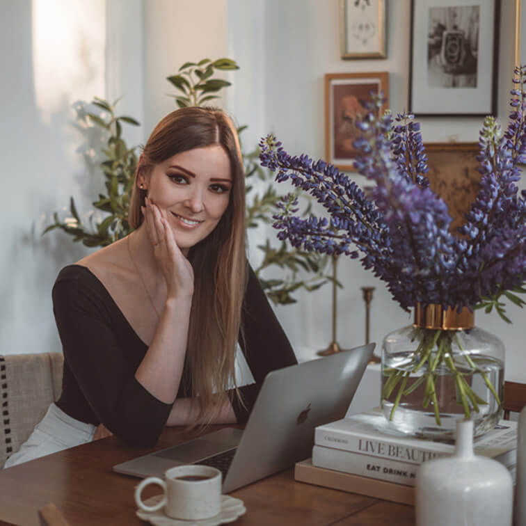 Frau sitzt an Holztisch mit Laptop und Glasvase mit violetten Blumen in hellem Raum