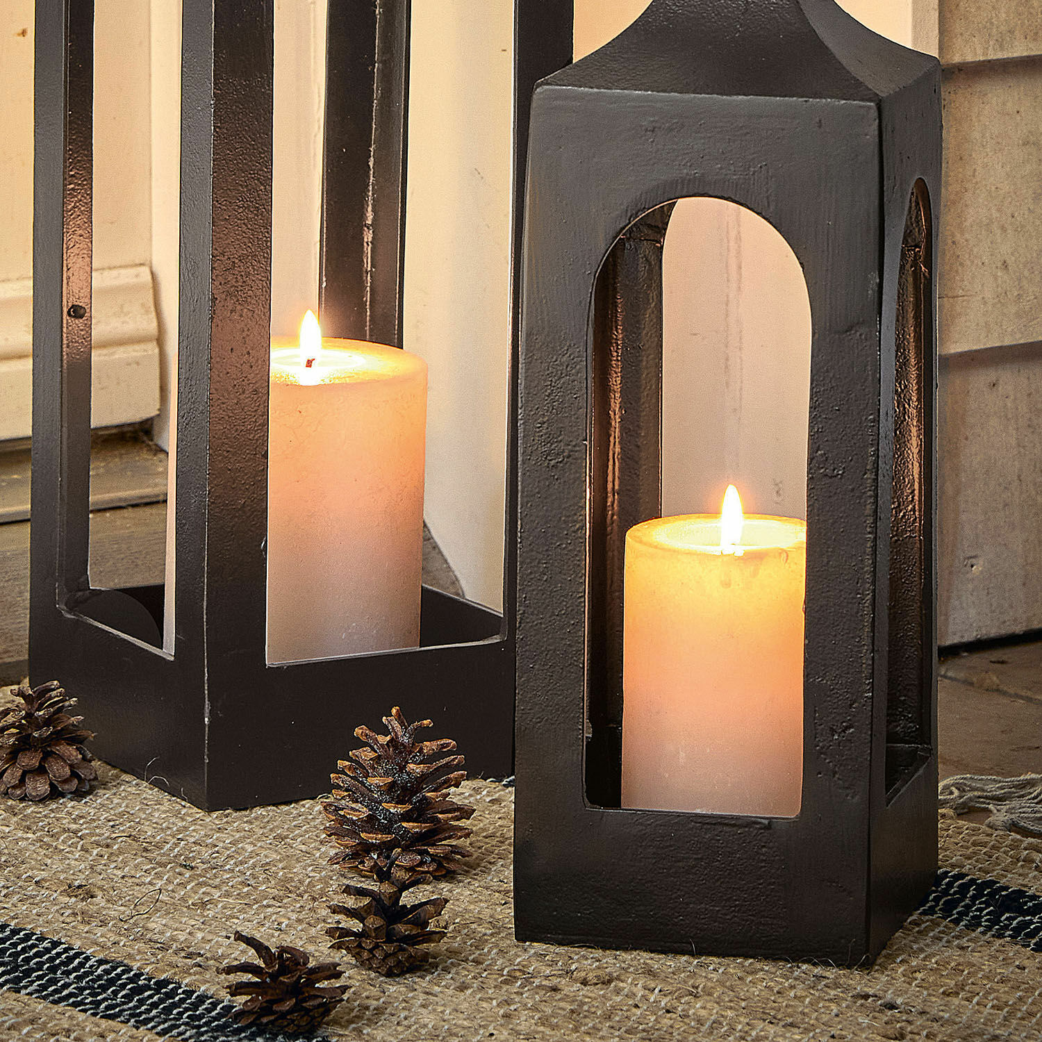 Schwarze Metalllaterne mit weißer Kerze und Kiefernzapfen auf Juteteppich, Wohnaccessoire