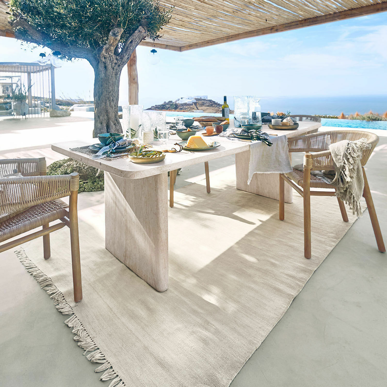 Heller Steintisch mit Naturteppich und Rattanstühlen auf Terrasse mit Meerblick