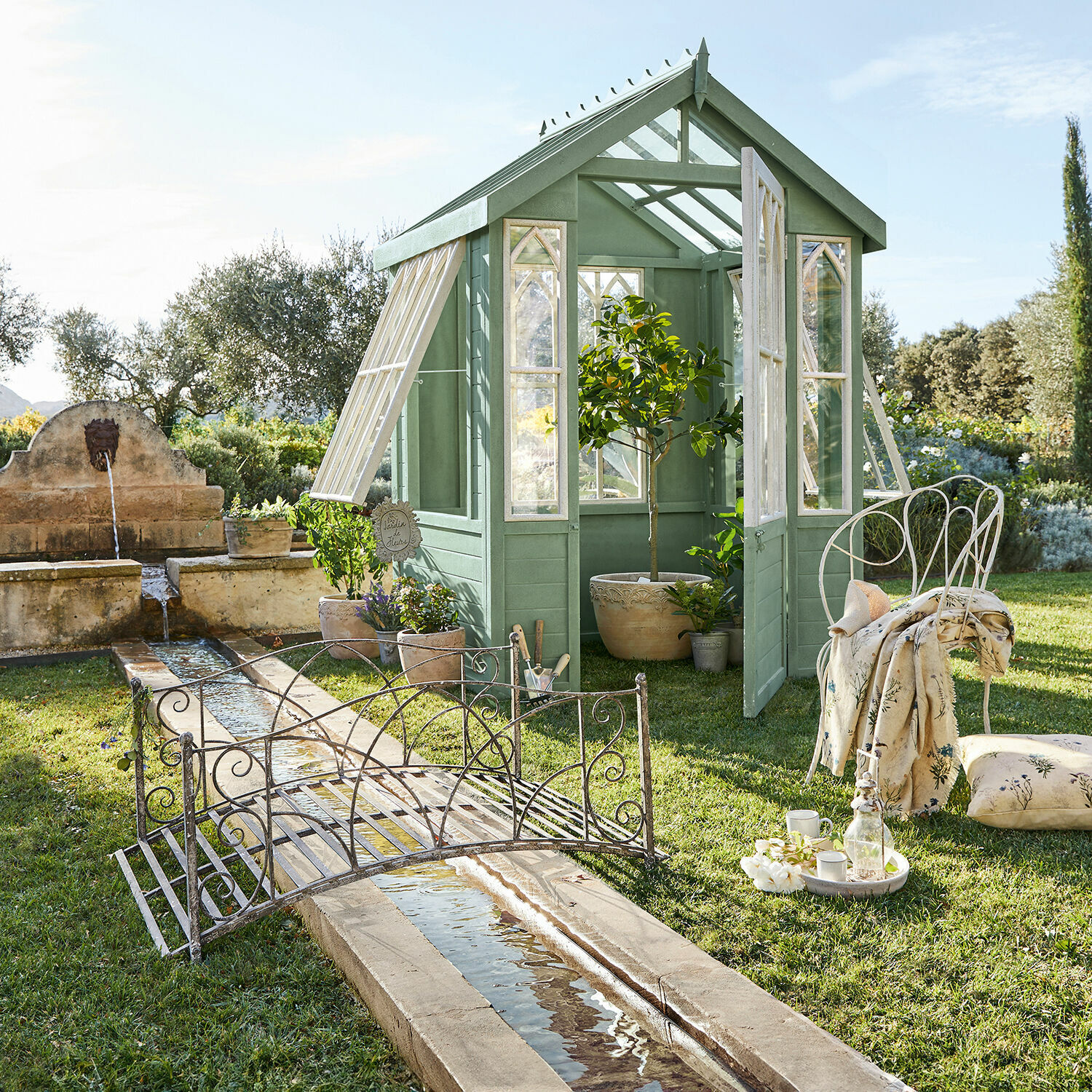 Grünes Holzgewächshaus im Garten mit dekorativem Metallbrückchen und Stuhl auf dem Rasen