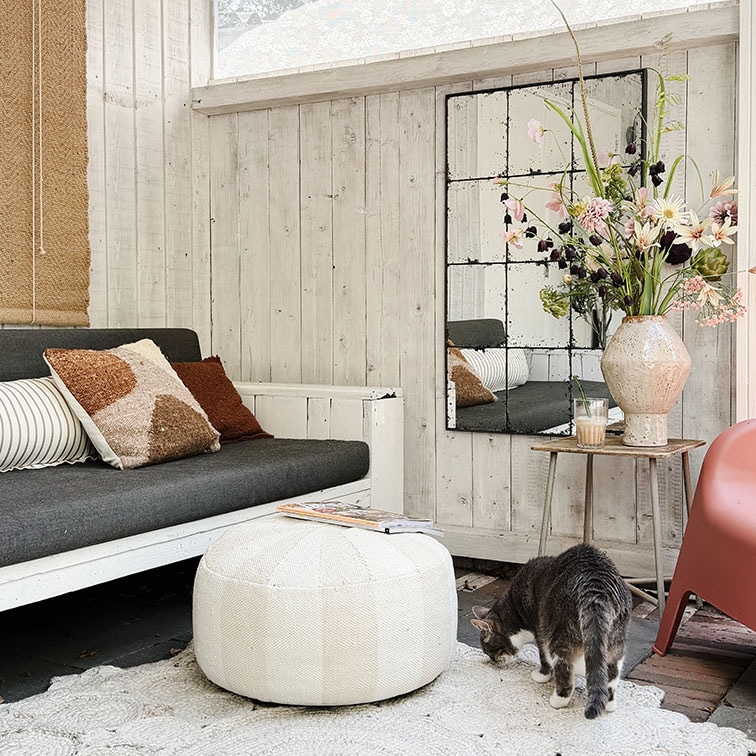 Gemütliches Wohnzimmer mit weißer Holzverkleidung, grauem Sofa, weißem Pouf und Blumenarrangement
