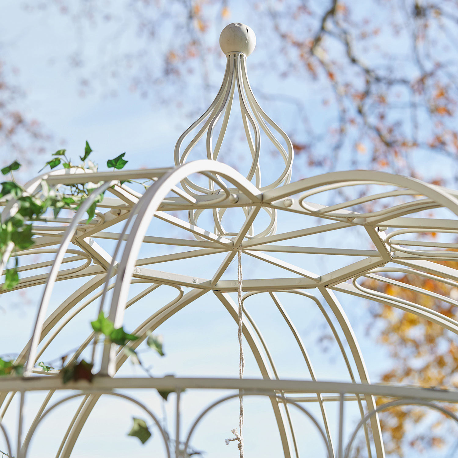 Dekorative cremefarbene Metallpergola mit filigranem Muster und Kletterpflanzen vor blauem Himmel
