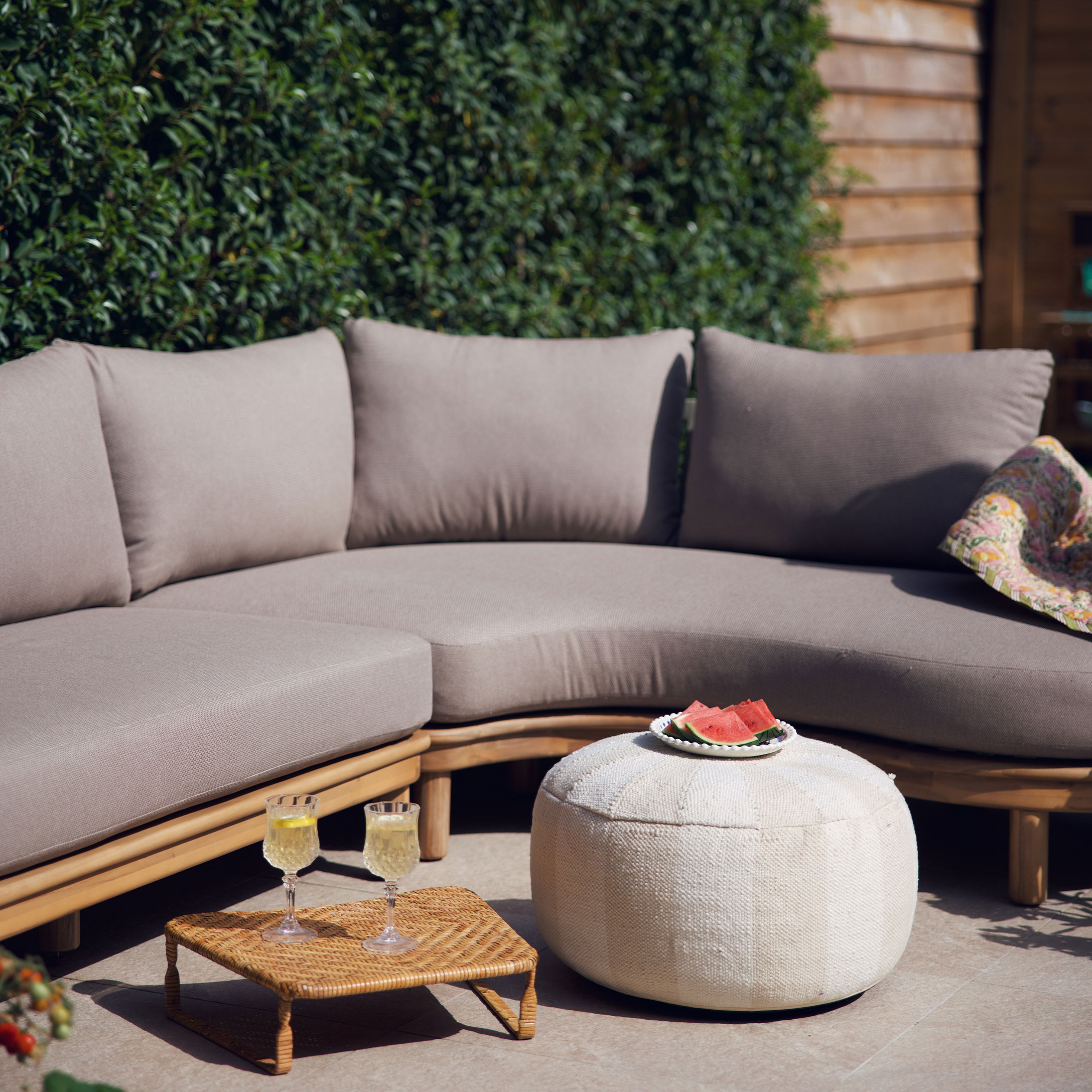 Beige Sofa mit Kissen auf Holzrahmen und weißer Pouf auf Terrasse mit sommerlicher Dekoration