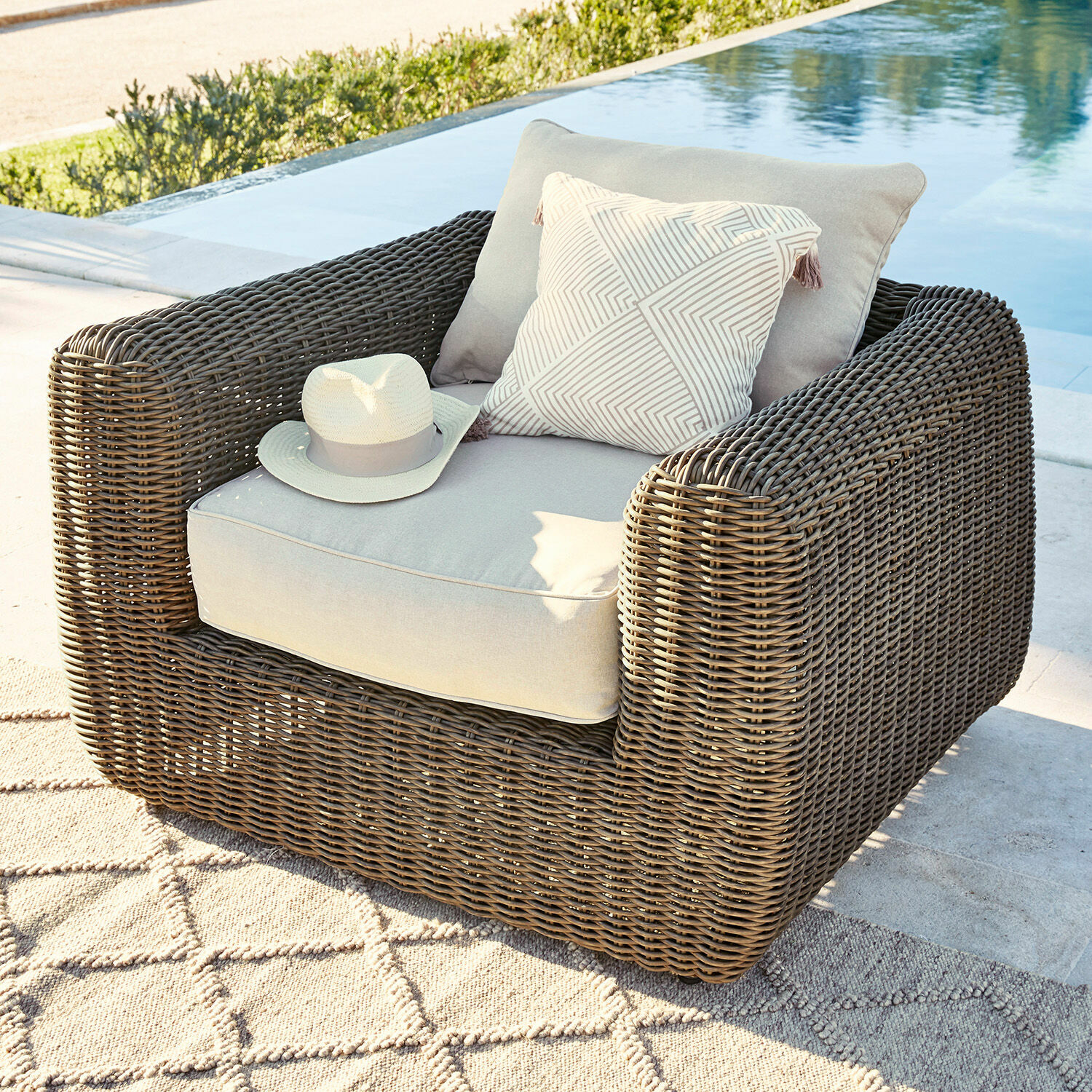 Sessel Bogdanne Gartenfauteuil aus braunem Rattan mit hellgrauen Polstern und Zierkissen am Pool auf der Terrasse