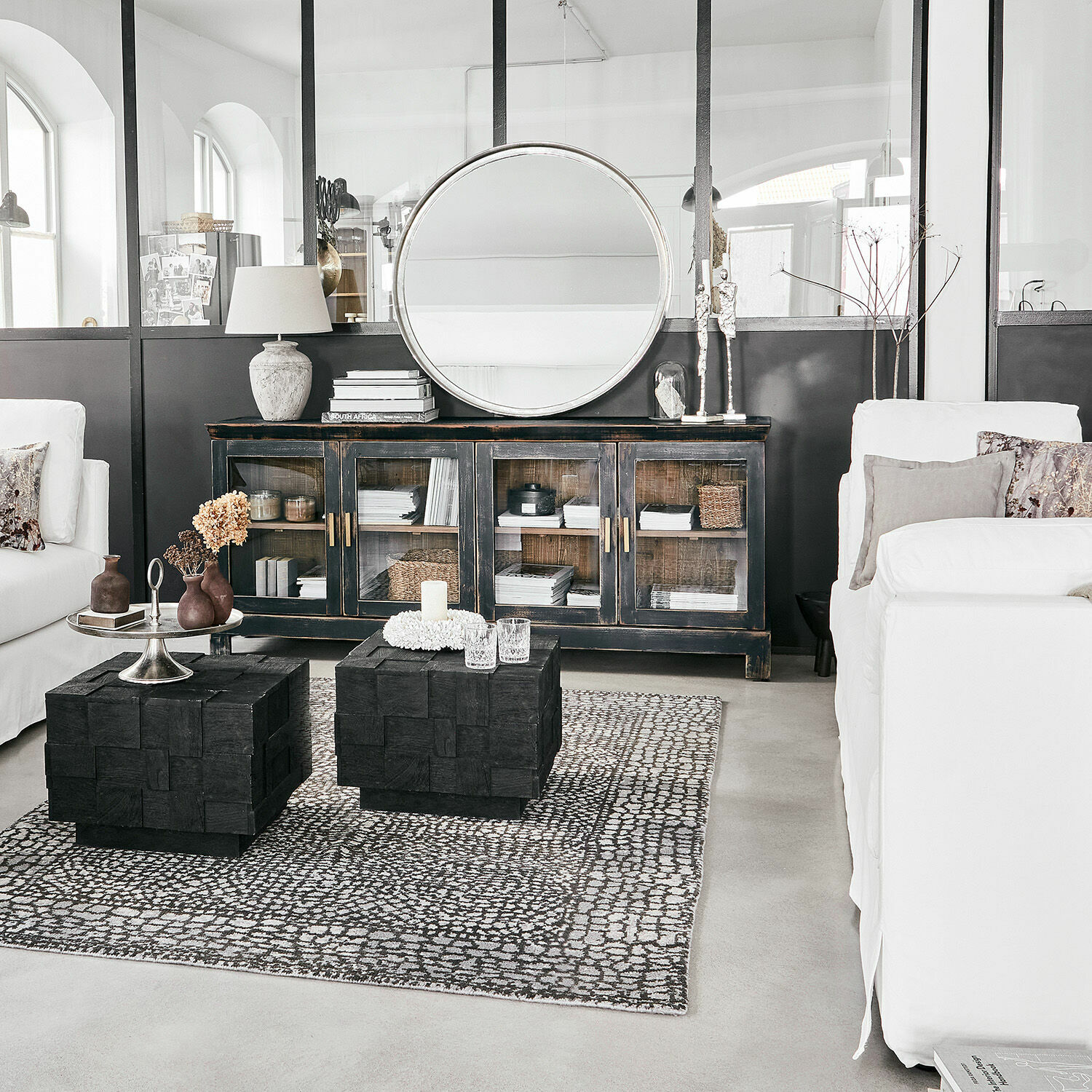 Wohnzimmer mit weißem Sofa, schwarzen Holztischen, Vitrine mit Glastüren und rundem Spiegel