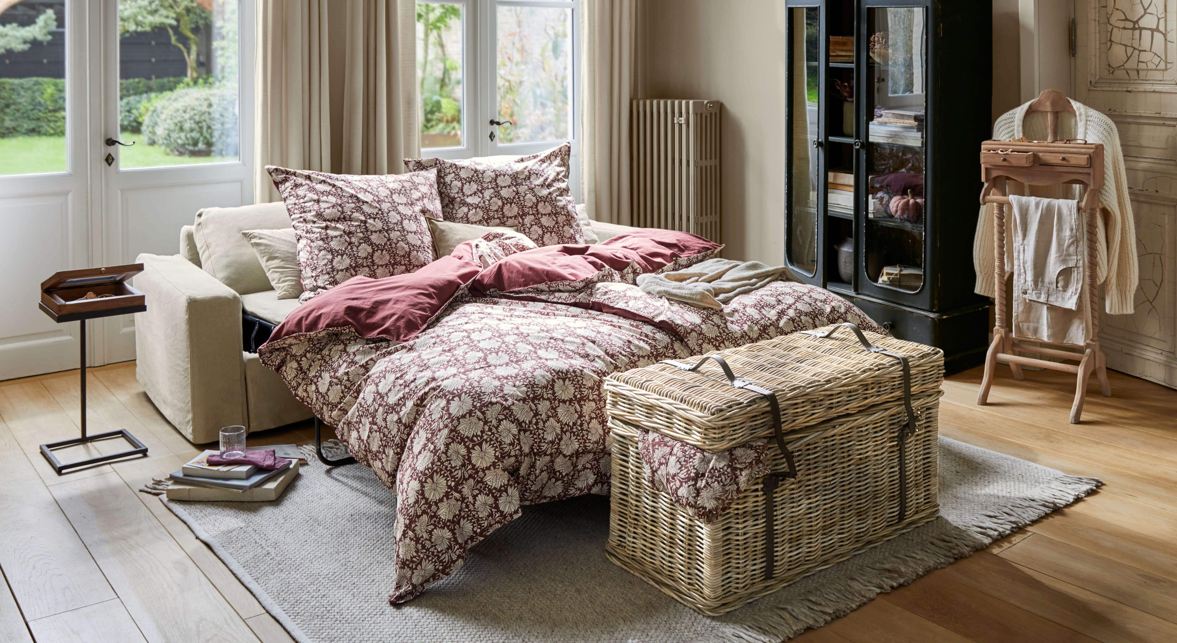 Ausziehbares Sofa mit beiger Polsterung und bordeauxfarbenem Bettzeug mit Blumenmuster im hellen Wohnzimmer