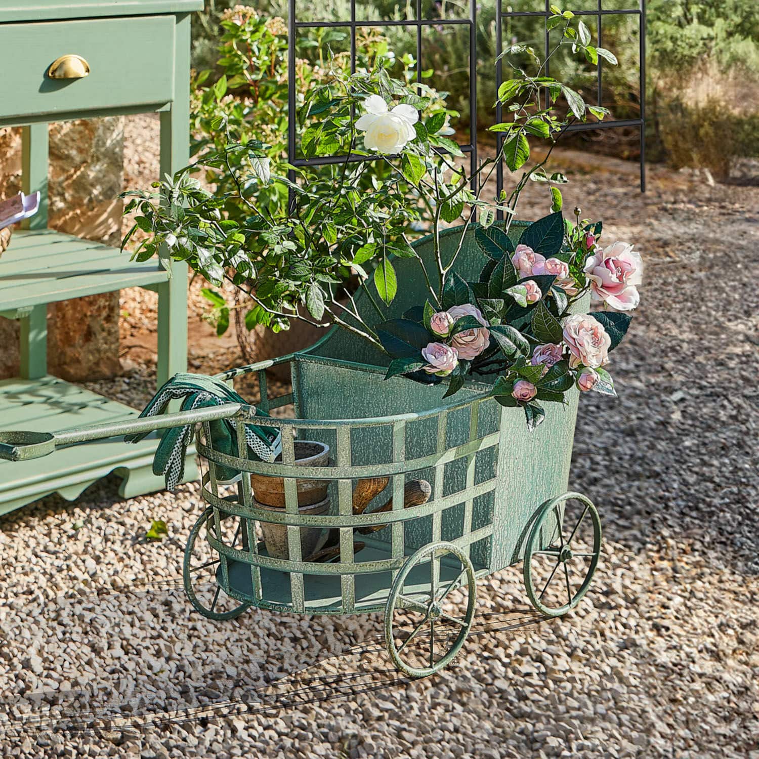 Grüner Metall-Gartenwagen mit Rosen und Blumentöpfen auf Kiesboden