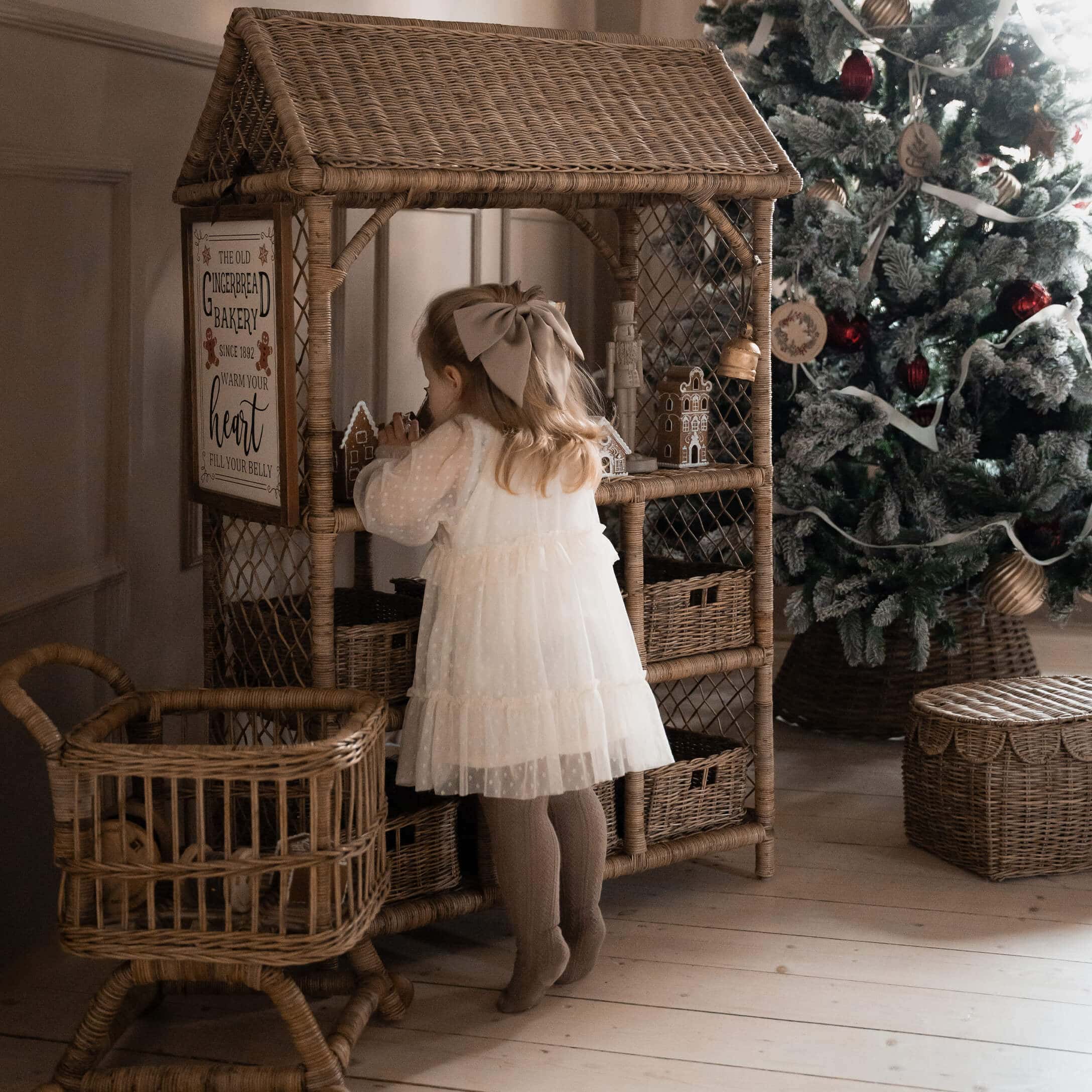 Kind in weißem Kleid vor einem Rattanschrank mit Lebkuchenhäusern neben geschmücktem Weihnachtsbaum