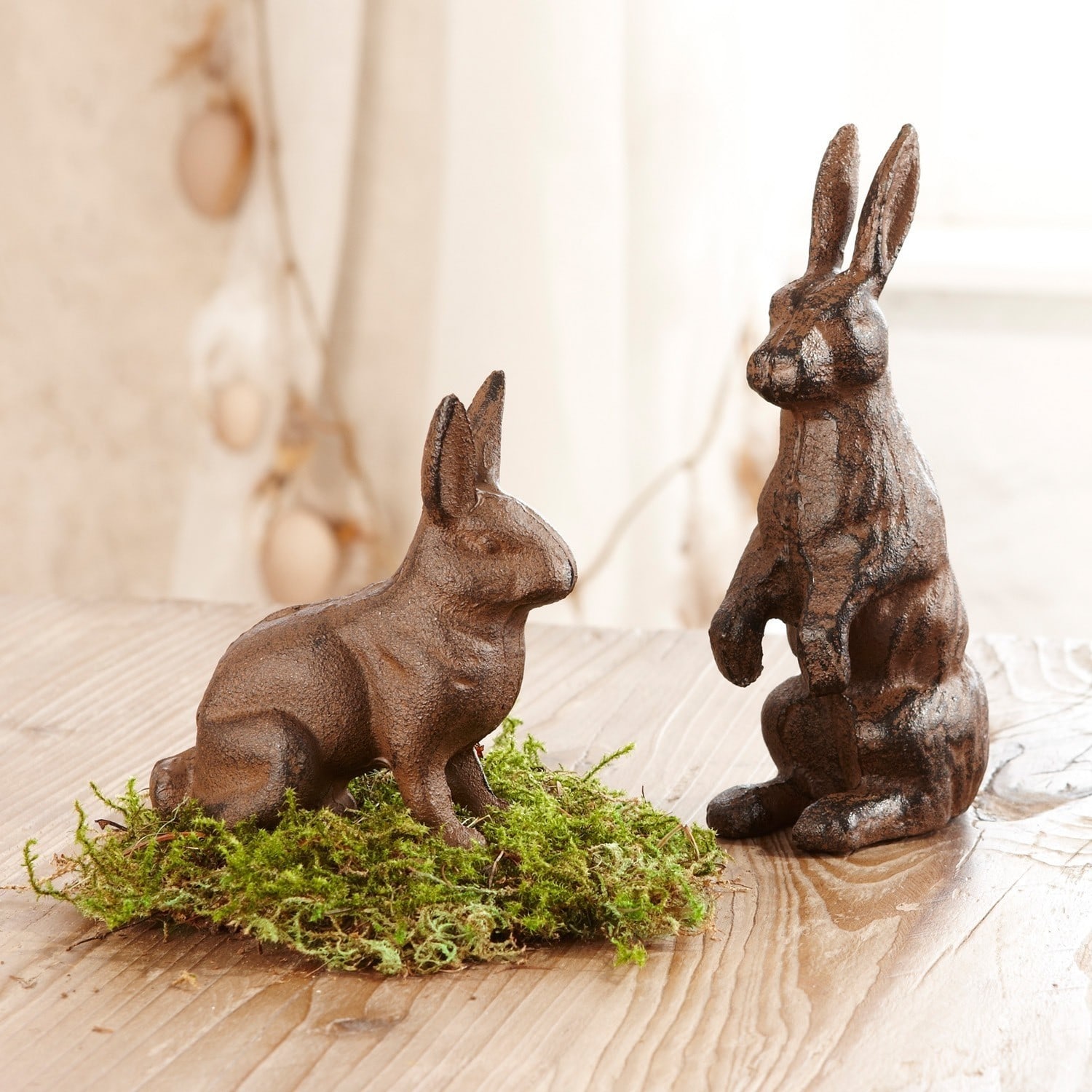 Zwei braune Metall-Hasenfiguren auf Holztisch mit grünem Moos als Dekoration