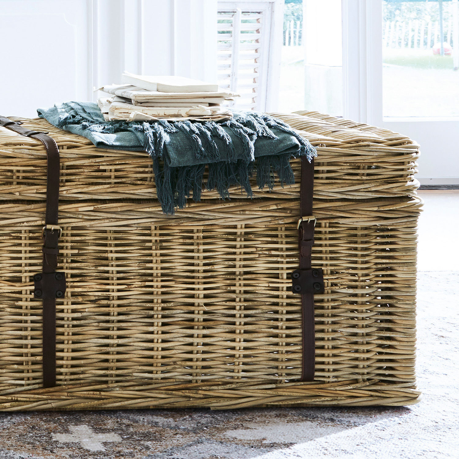 Große Rattankiste mit braunen Riemen und blauem Fransenplaid auf grauem Teppich