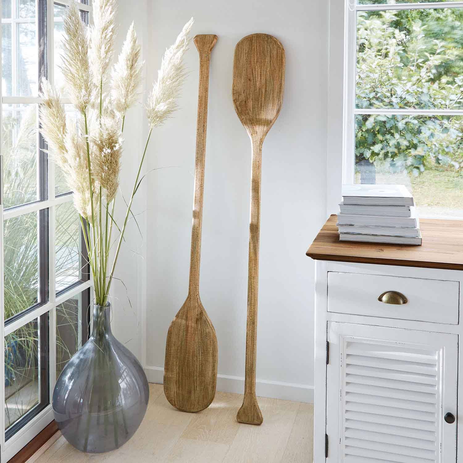 Zwei dekorative Paddel aus Holz in Naturfarbe lehnen an weißer Wand in hellem Raum