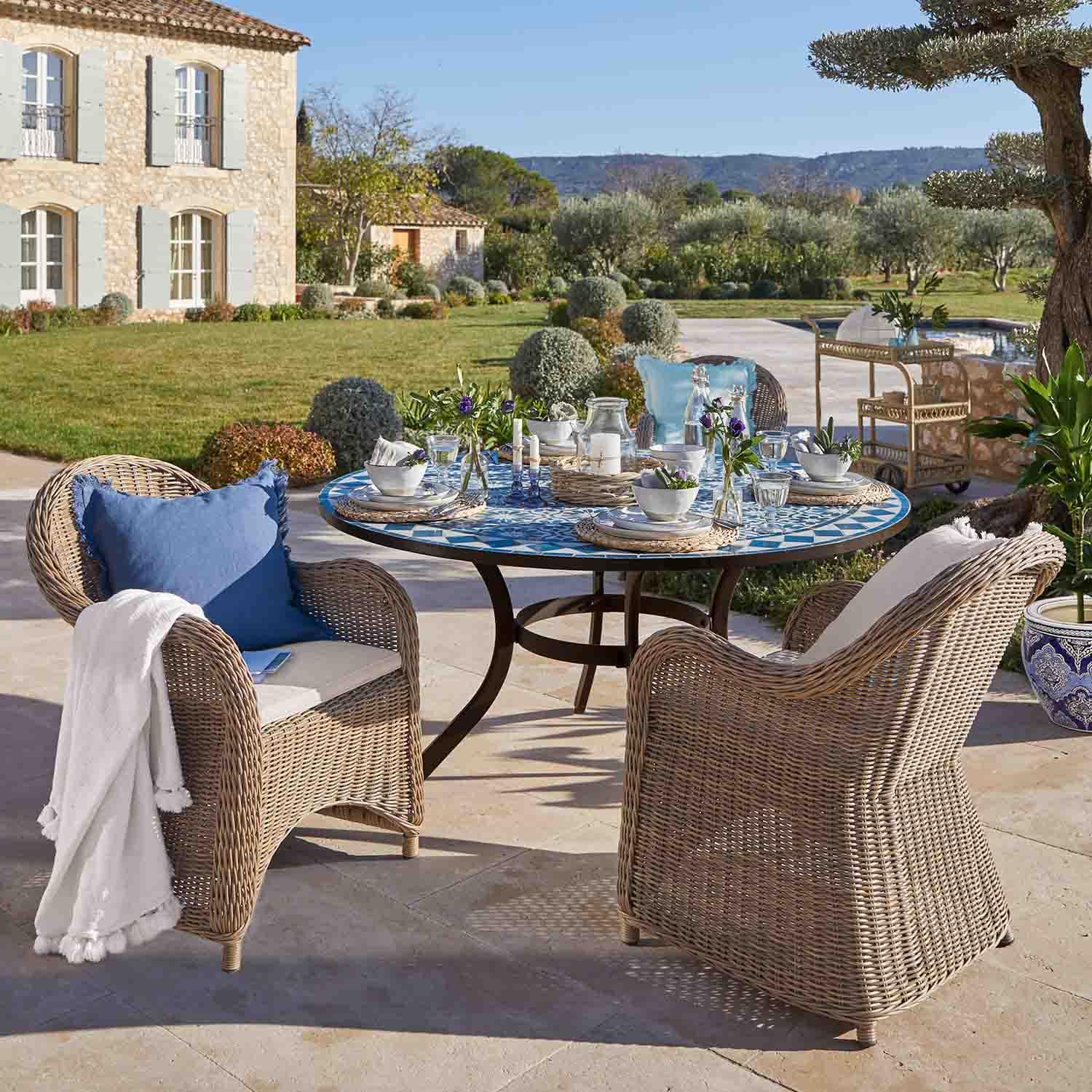 Runder Mosaiktisch in Blau mit beigen Rattanstühlen auf sonniger Gartenterrasse