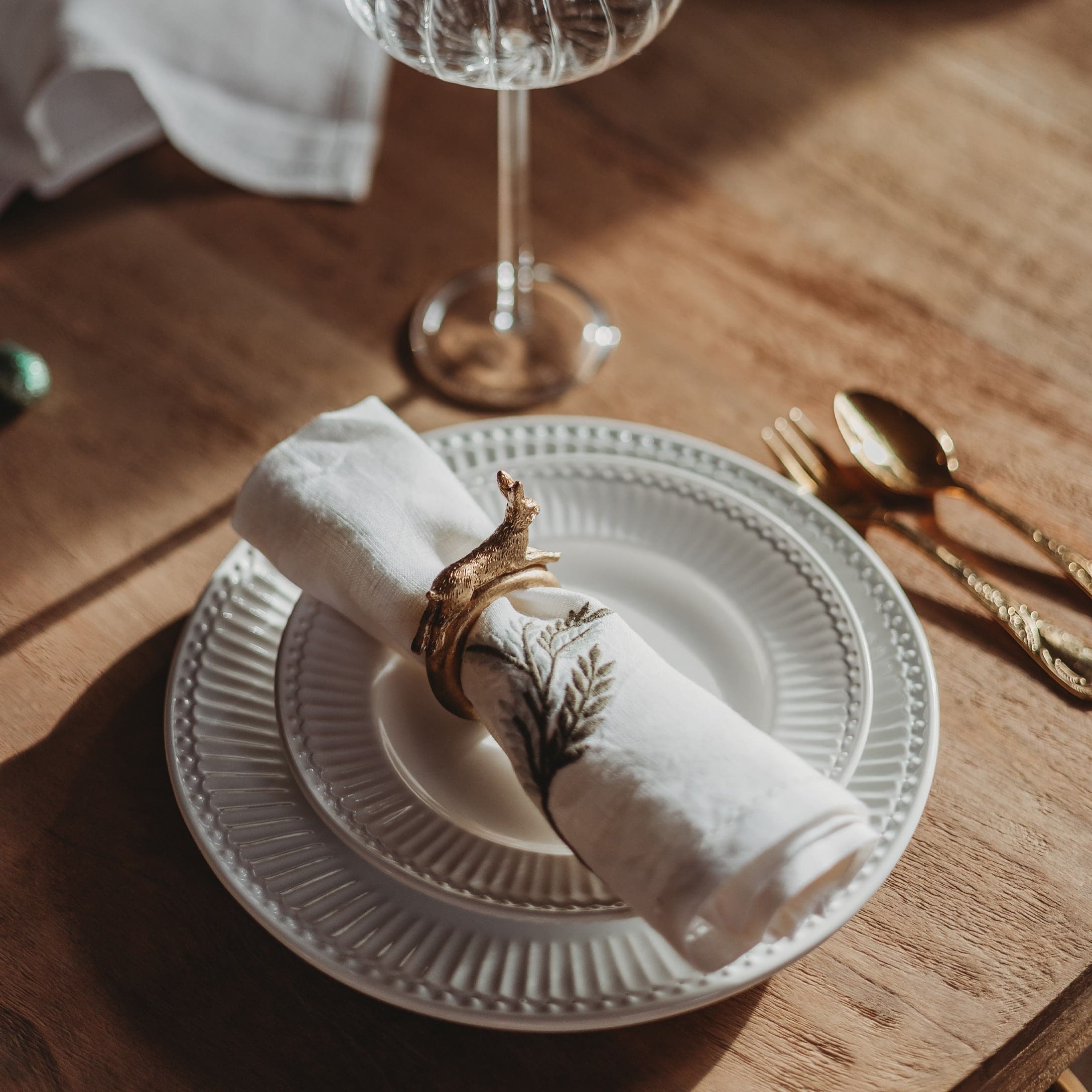 Weißer Keramikteller mit bestickter Serviette und goldenem Serviettenring auf Holztisch