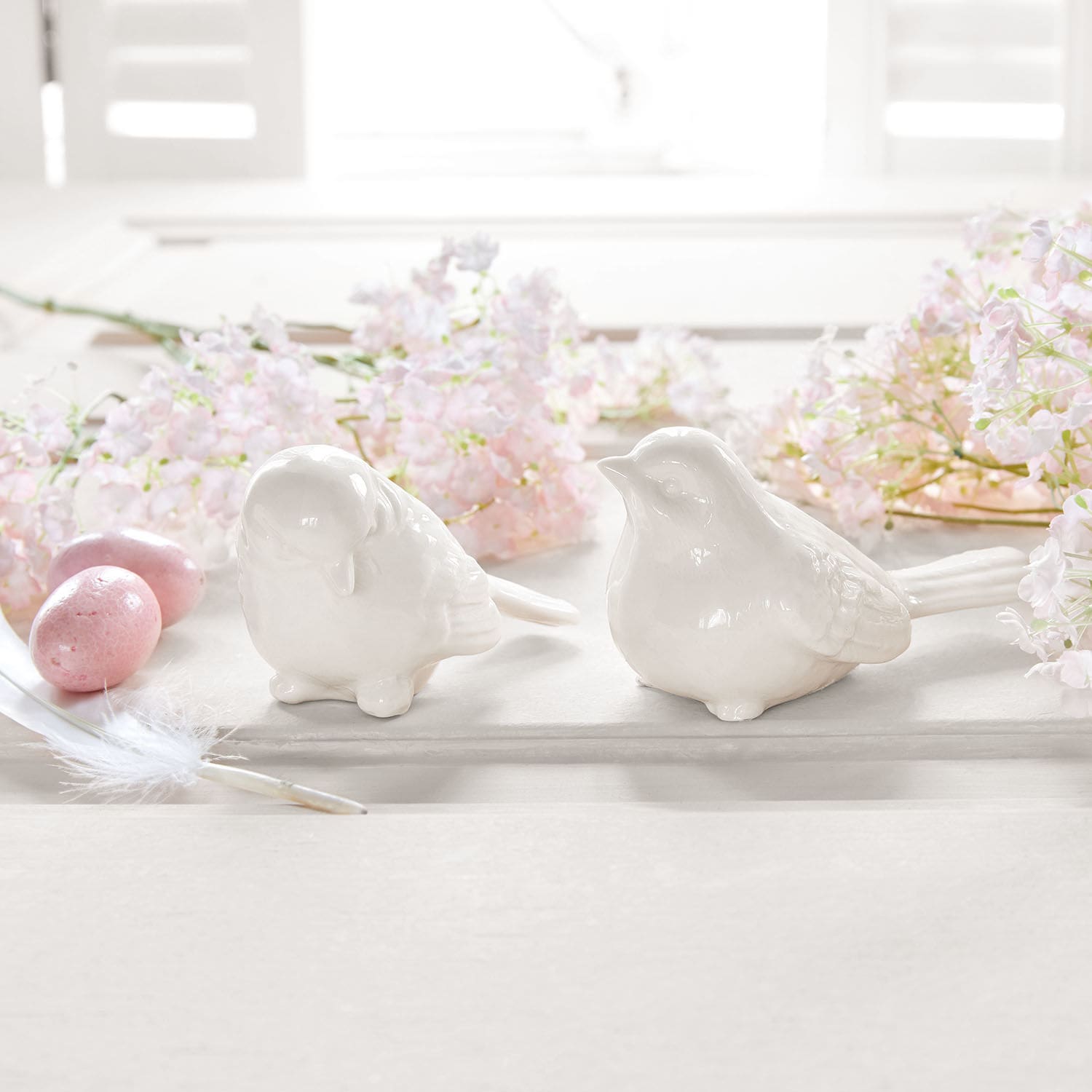 Zwei weiße Keramikvogel-Figuren auf hellem Untergrund mit rosa Blumen und Dekoeiern