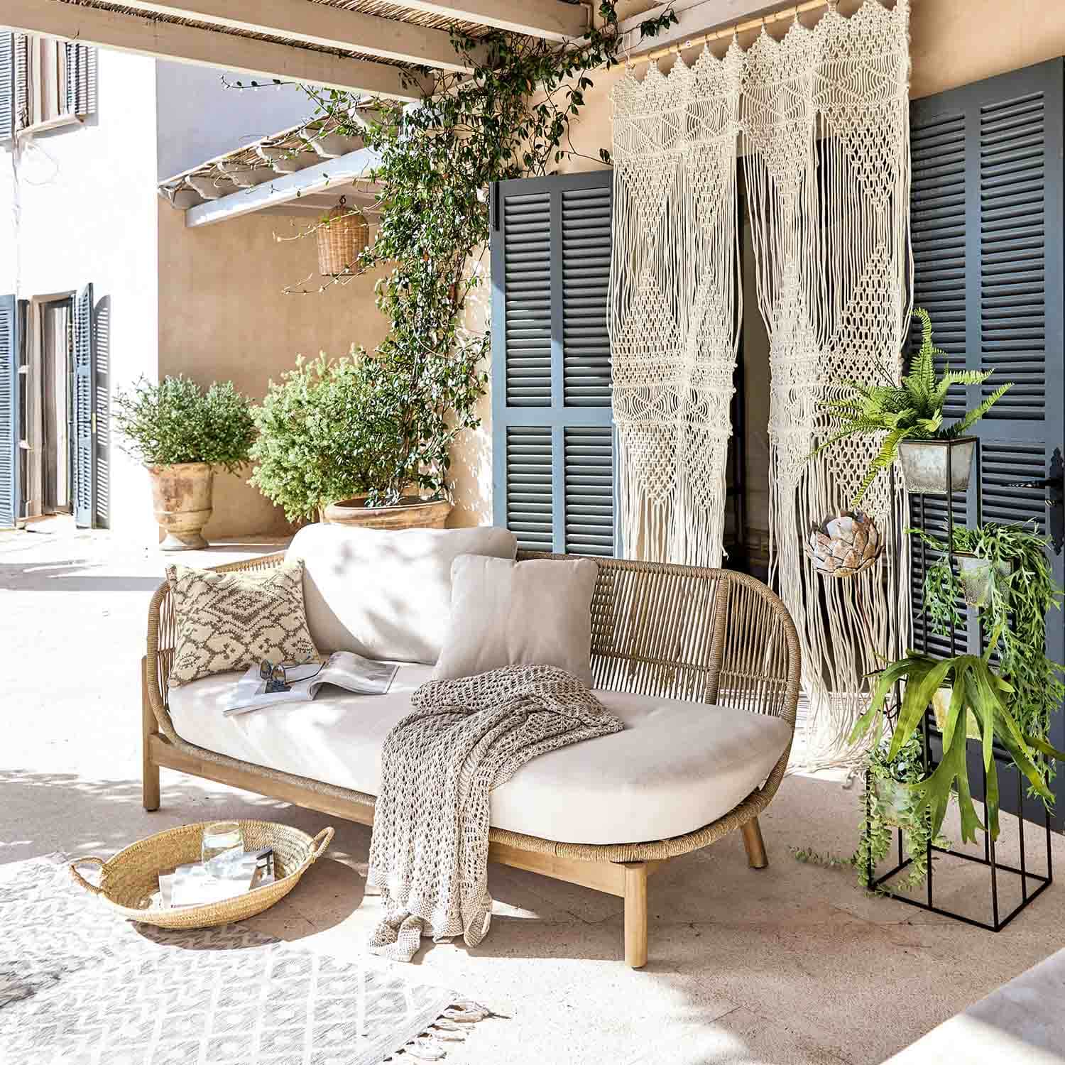 Rattansofa mit cremefarbenen Kissen und gestrickter Decke auf heller Terrasse mit Pflanzen